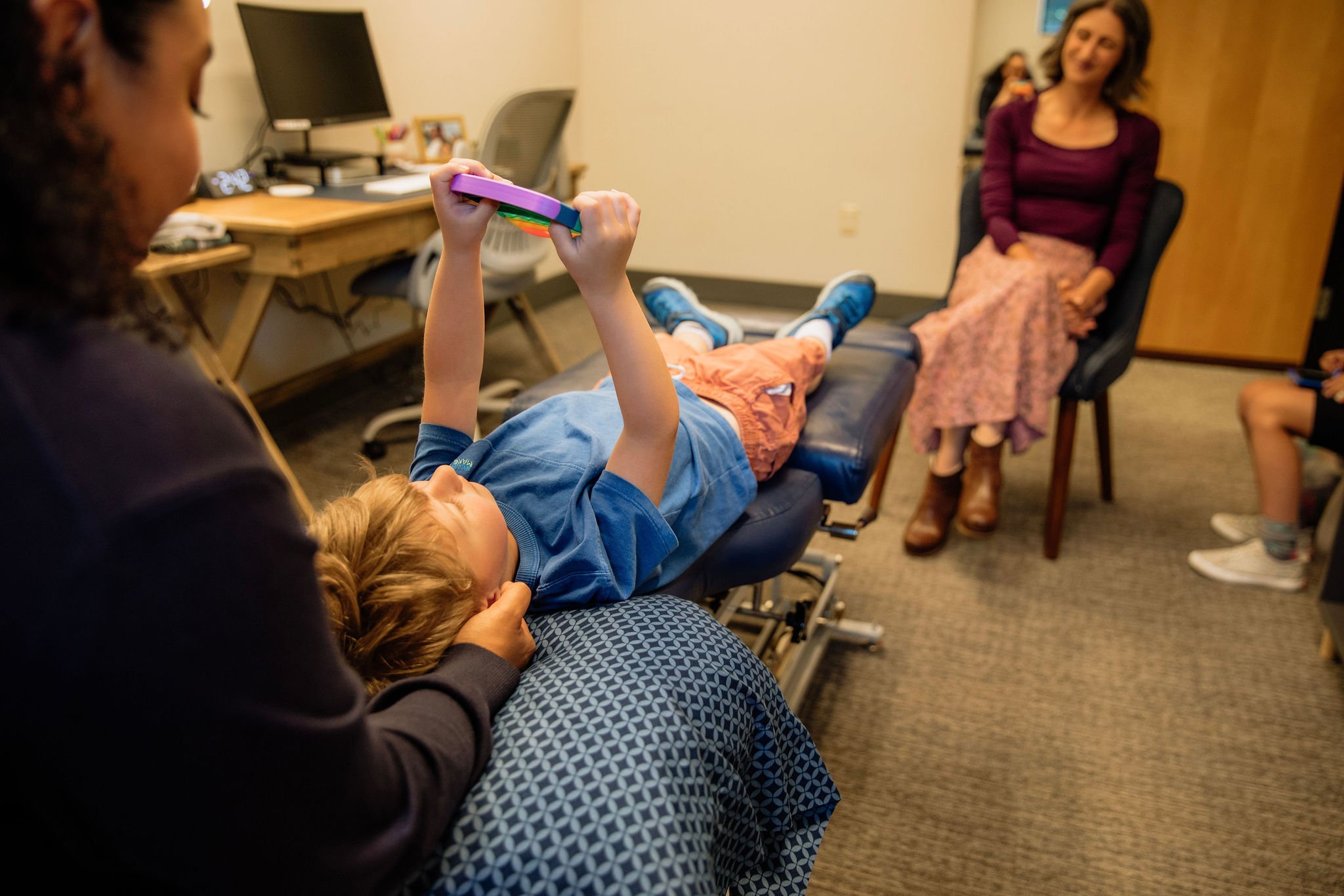 A young boy lying on a medical examination table with his legs and head supported, holding a colorful stack of paper plates, while a woman in a maroon top and floral skirt sits nearby and smiles. A woman with dark hair is partially visible on the left, and another person is on the right. The room appears to be an office with a desk, computer, and personal items.