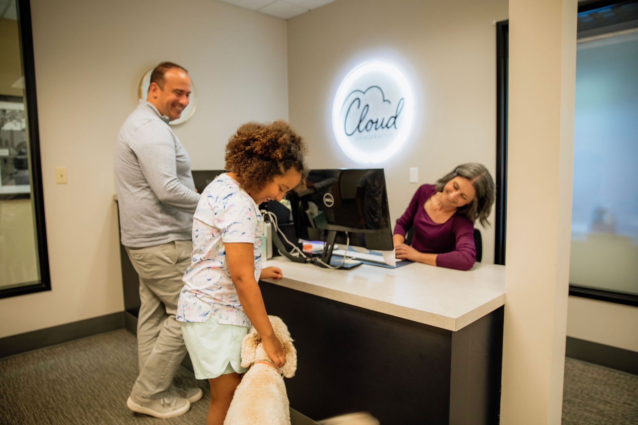 A woman with curly hair at a reception desk holding a large dog, with two smiling people behind her, in an office or clinic setting with a sign that says 'Cloud' on the wall.