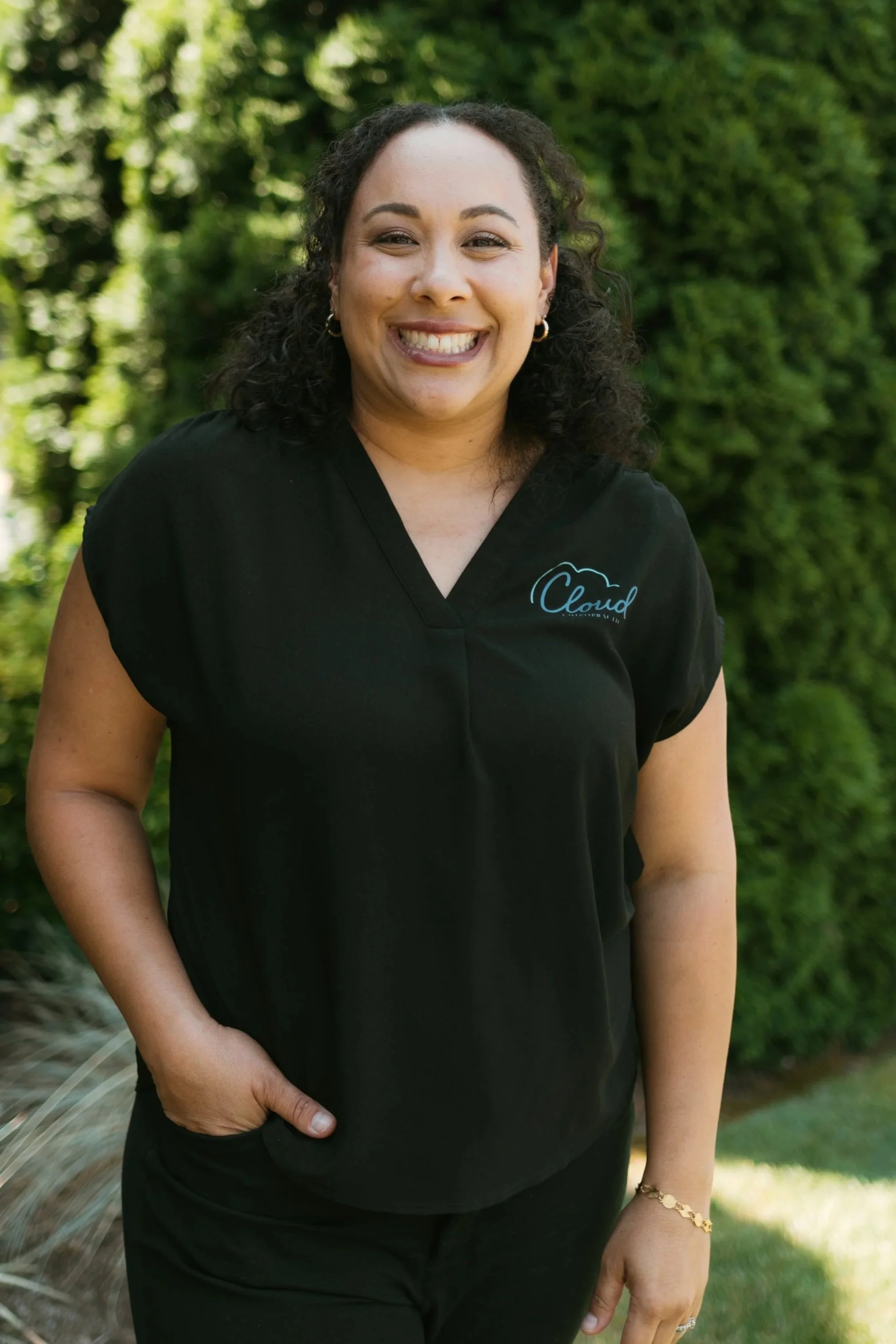 A woman with dark curly hair smiling outdoors in front of green trees, wearing a black shirt with a logo that says 'Cloud' and a gold bracelet.
