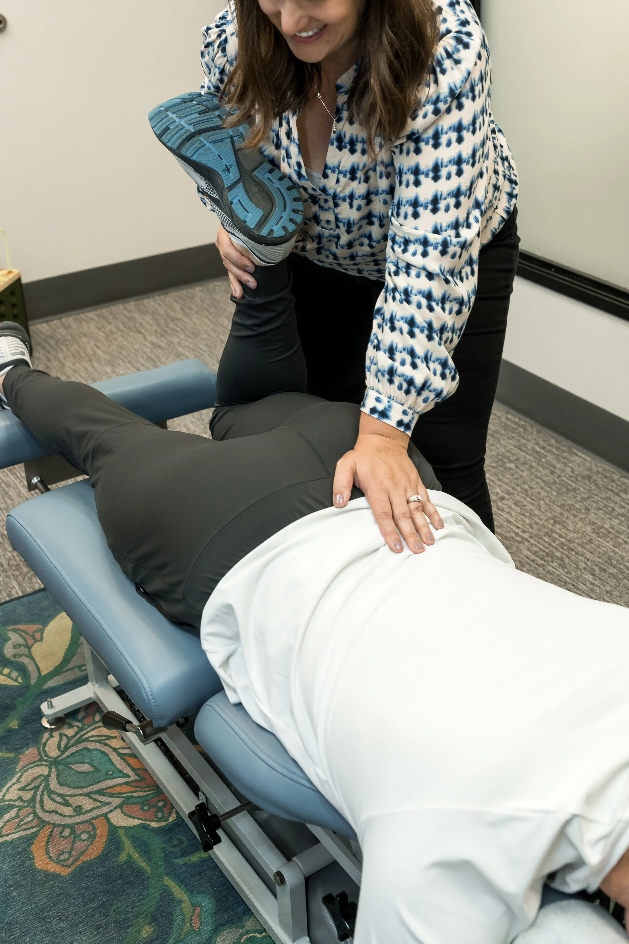 A woman dressed in a blue and white patterned blouse is assisting a person lying face down on a massage or therapy table, holding their leg upright during a physical therapy or chiropractic session.