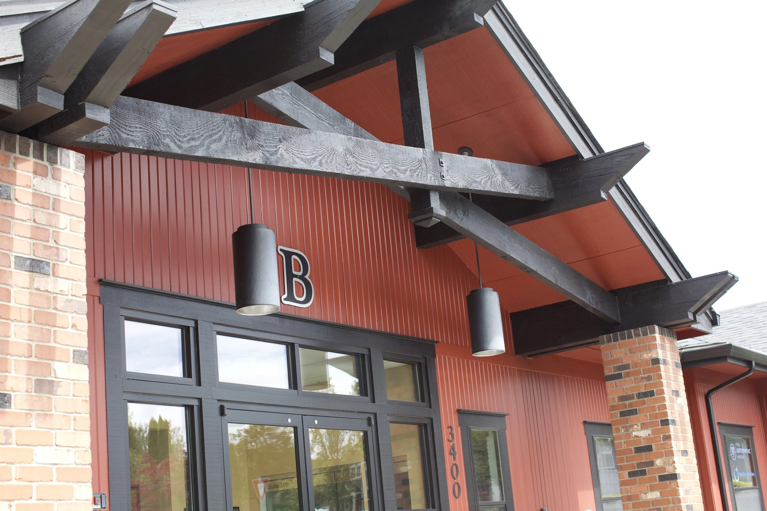 Exterior view of a modern commercial building with red vertical siding, brick accents, large windows, and a prominent wooden canopy structure above the entrance, with hanging lights.