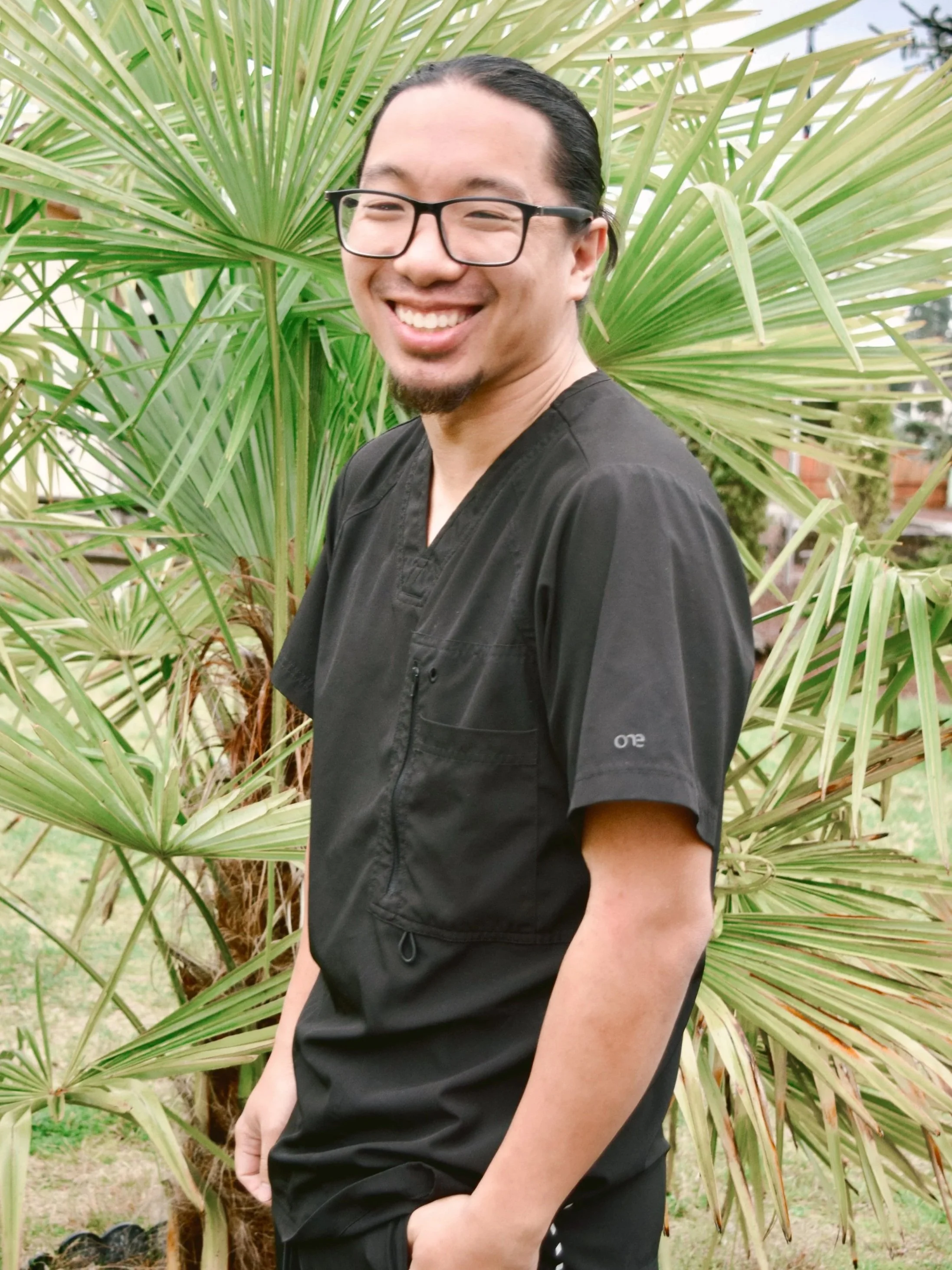 A man smiling in front of a green palm tree, wearing black scrubs and glasses.