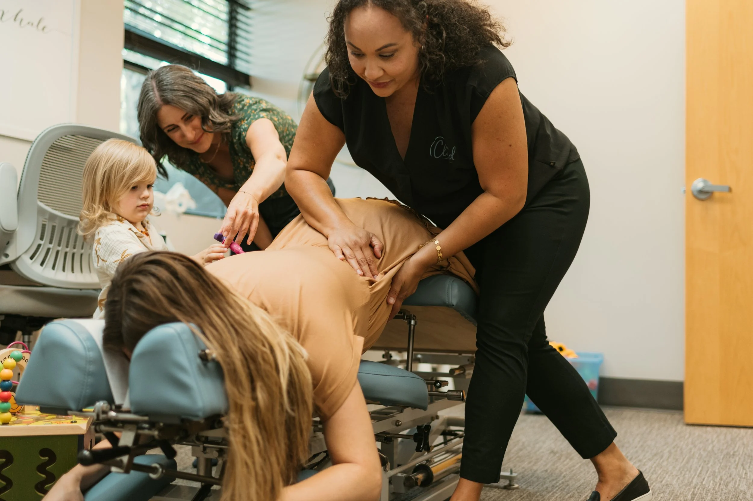 A group of children and two women in a therapy or medical setting. One woman is lying on her stomach on a treatment table, while another woman is applying pressure to her back. The children are nearby, observing the procedure.