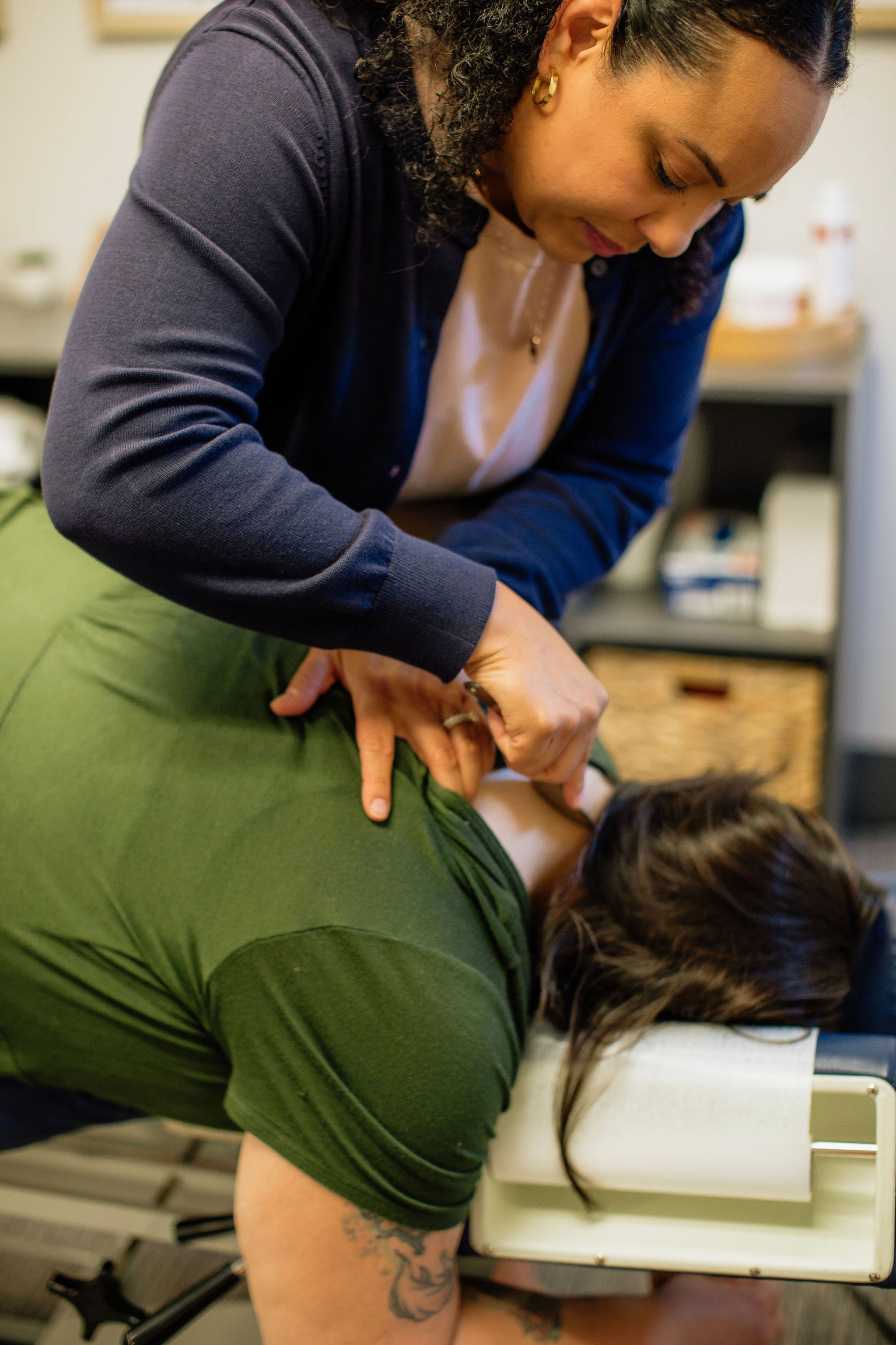 Two women administering a medical or healthcare procedure, one lying on a table and the other leaning over her, in a clinical setting.