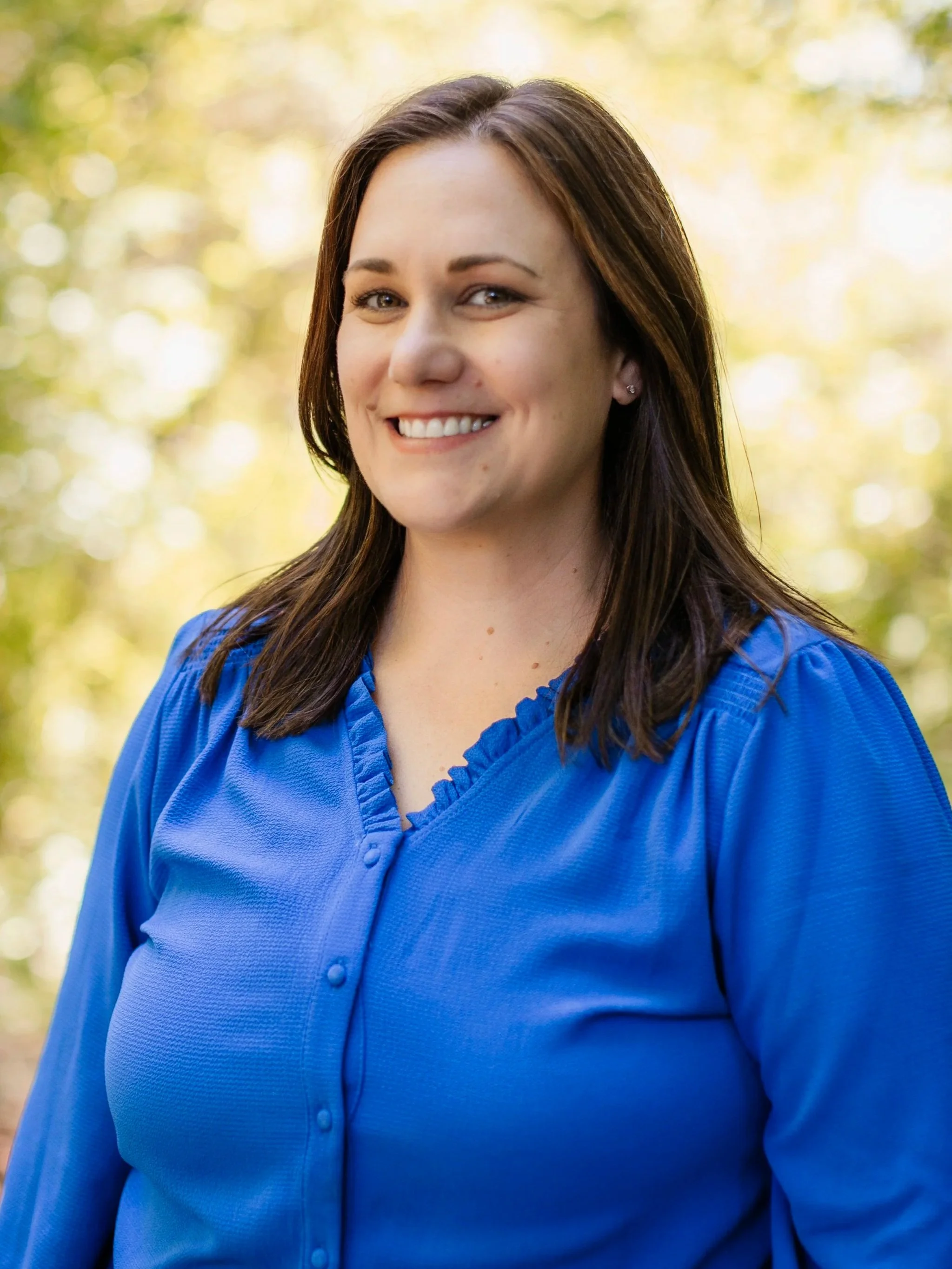 Close-up of a woman with brown hair and blue eyes smiling outdoors, wearing a blue shirt with a ruffled collar.