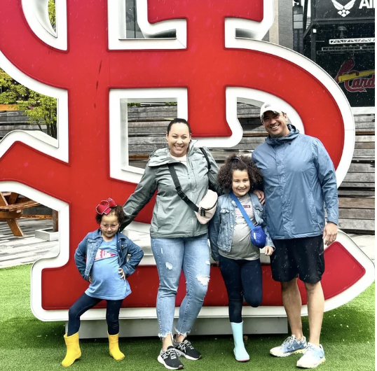 Family of four standing in front of a large red and white letters sign, outdoors, smiling, with green grass and wooden structures in the background.