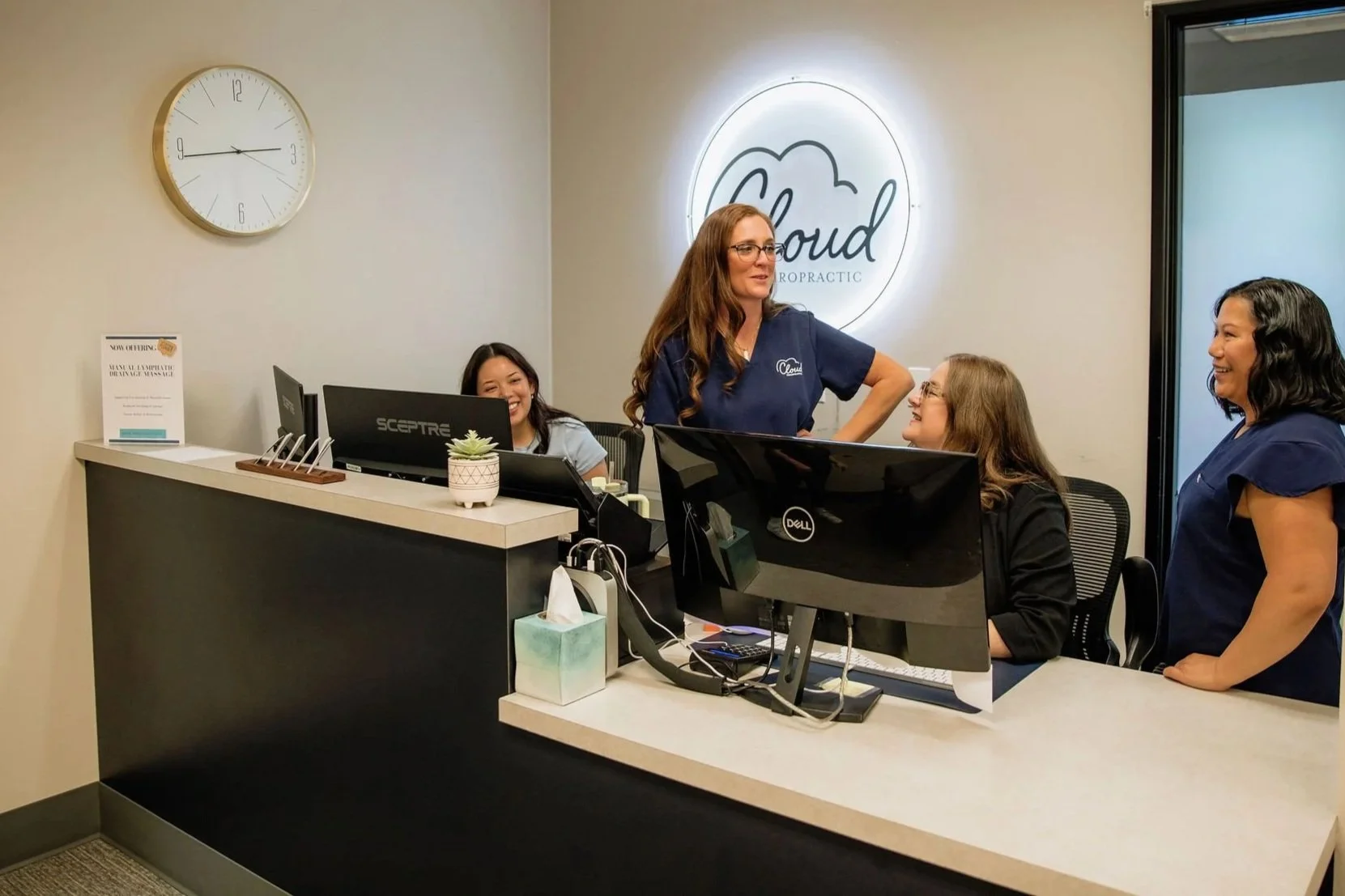 Four women inside a chiropractic office reception area, two standing and two sitting. They are smiling and engaging in conversation. The office has a modern look with a reception desk, a plant, a clock, and a neon sign that reads 'Cloud Chiropractic' on the back wall.