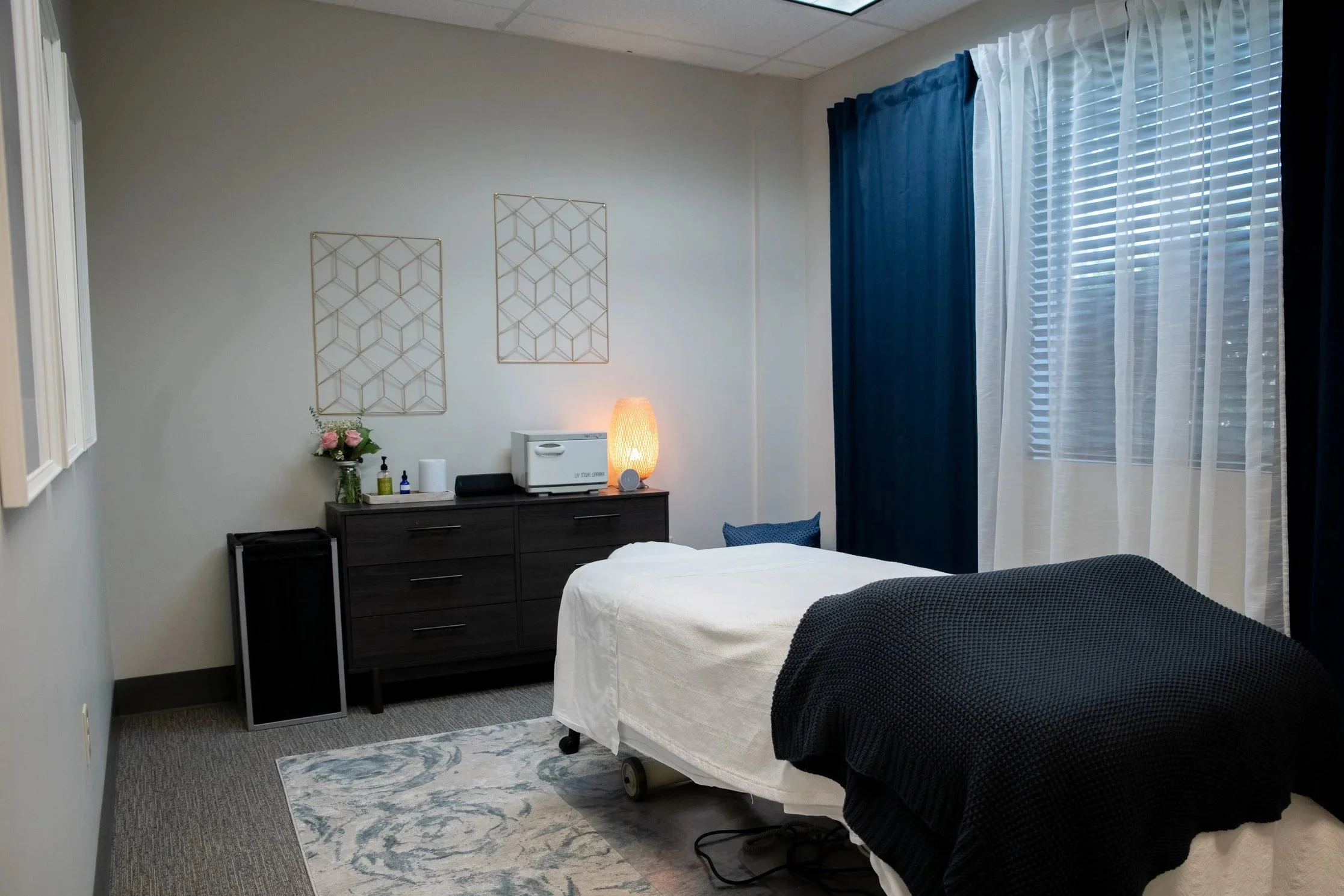 A hotel room with a bed, a dresser with a lamp, flowers, and personal items, and large windows with blue curtains and white sheer curtains.