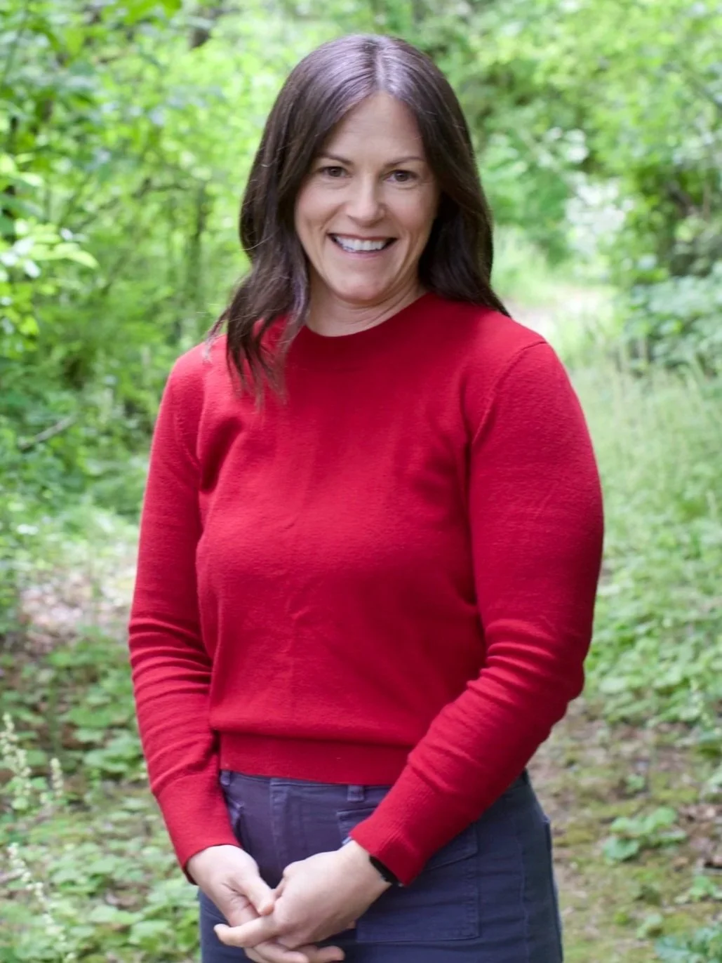 A woman with shoulder-length dark hair smiling outdoors in a green forest, wearing a red sweater and dark pants.