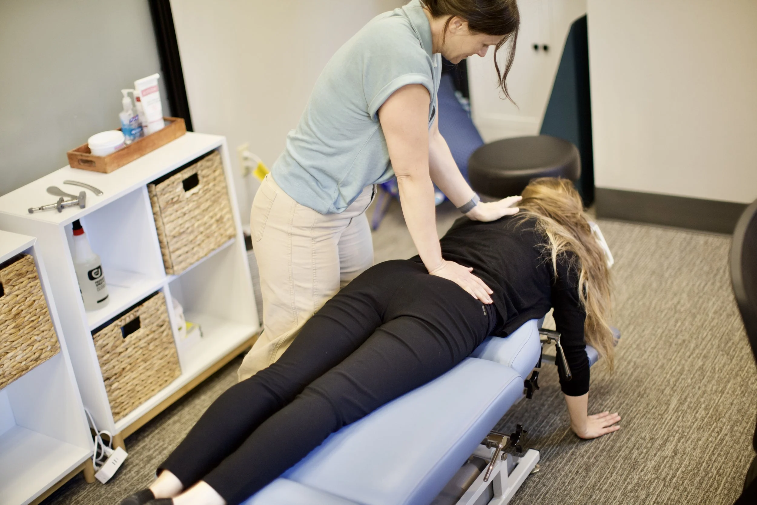 A physical therapist guides a woman performing a stretching exercise on a treatment table in a clinical setting.