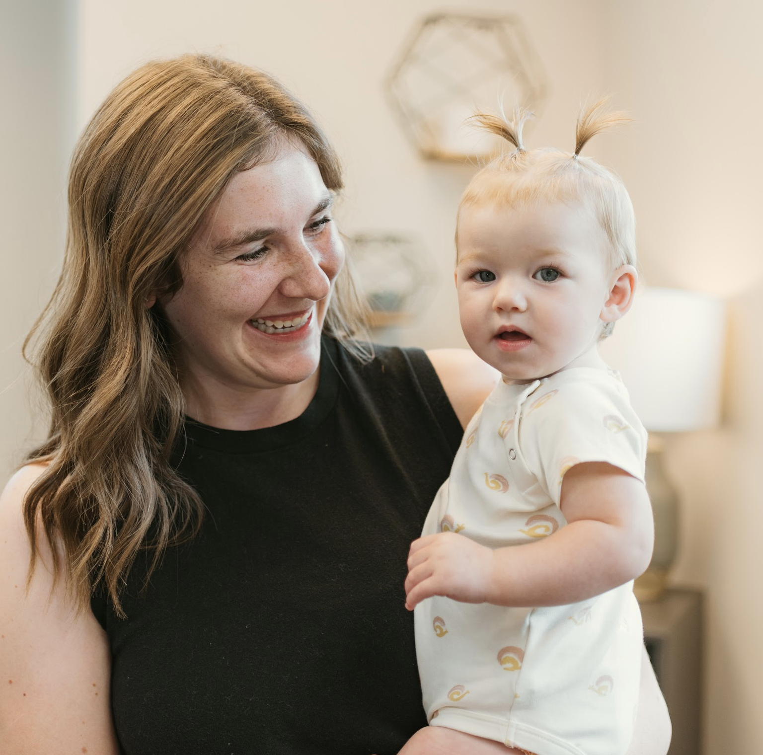 A woman with long, brown hair smiling and holding a young girl with blond hair styled in two small ponytails. The girl is wearing a light-colored romper with a rainbow pattern.