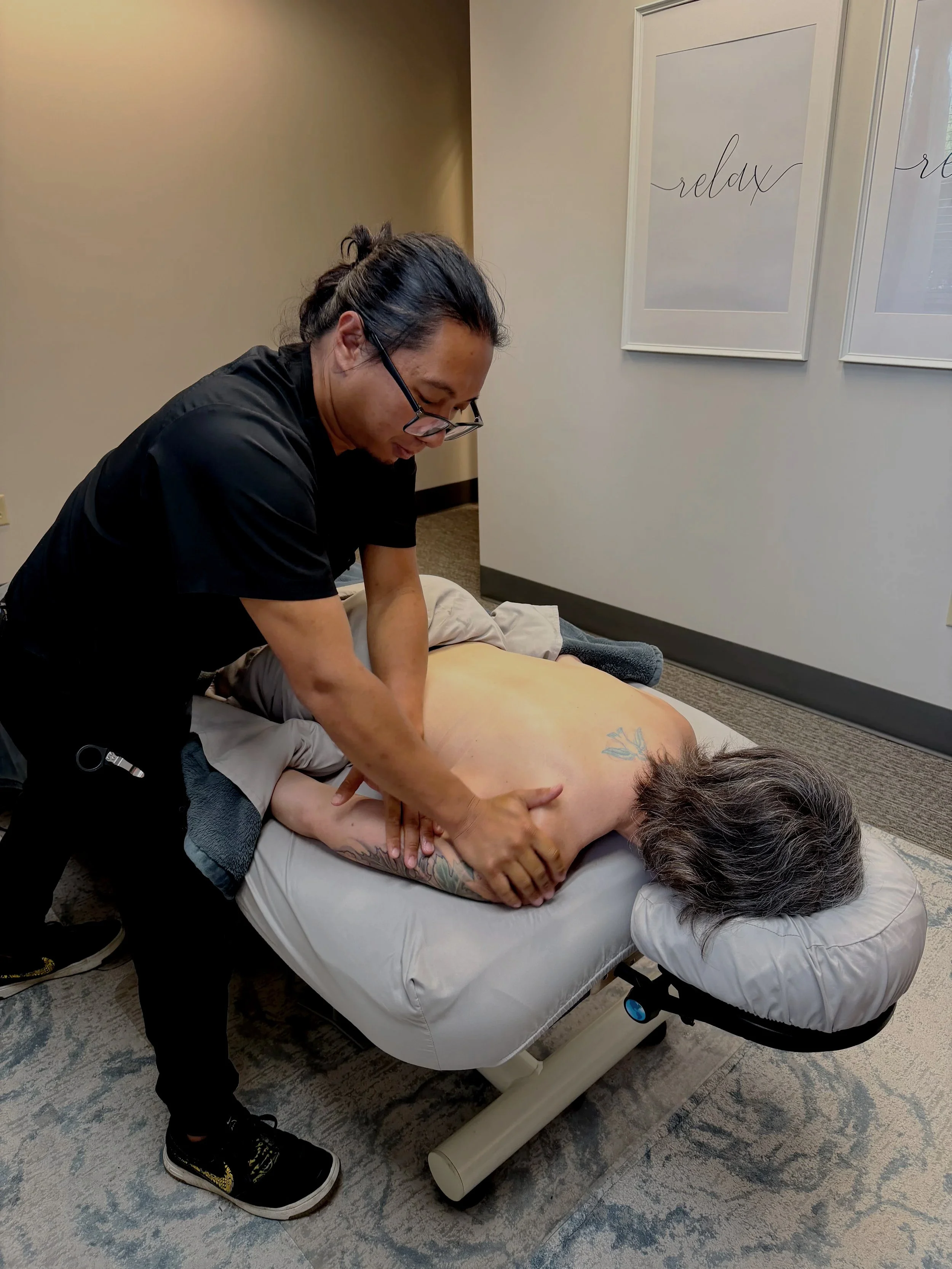A person receiving a back massage from a massage therapist on a massage table in a room with framed artwork on the wall that reads 'relax'.