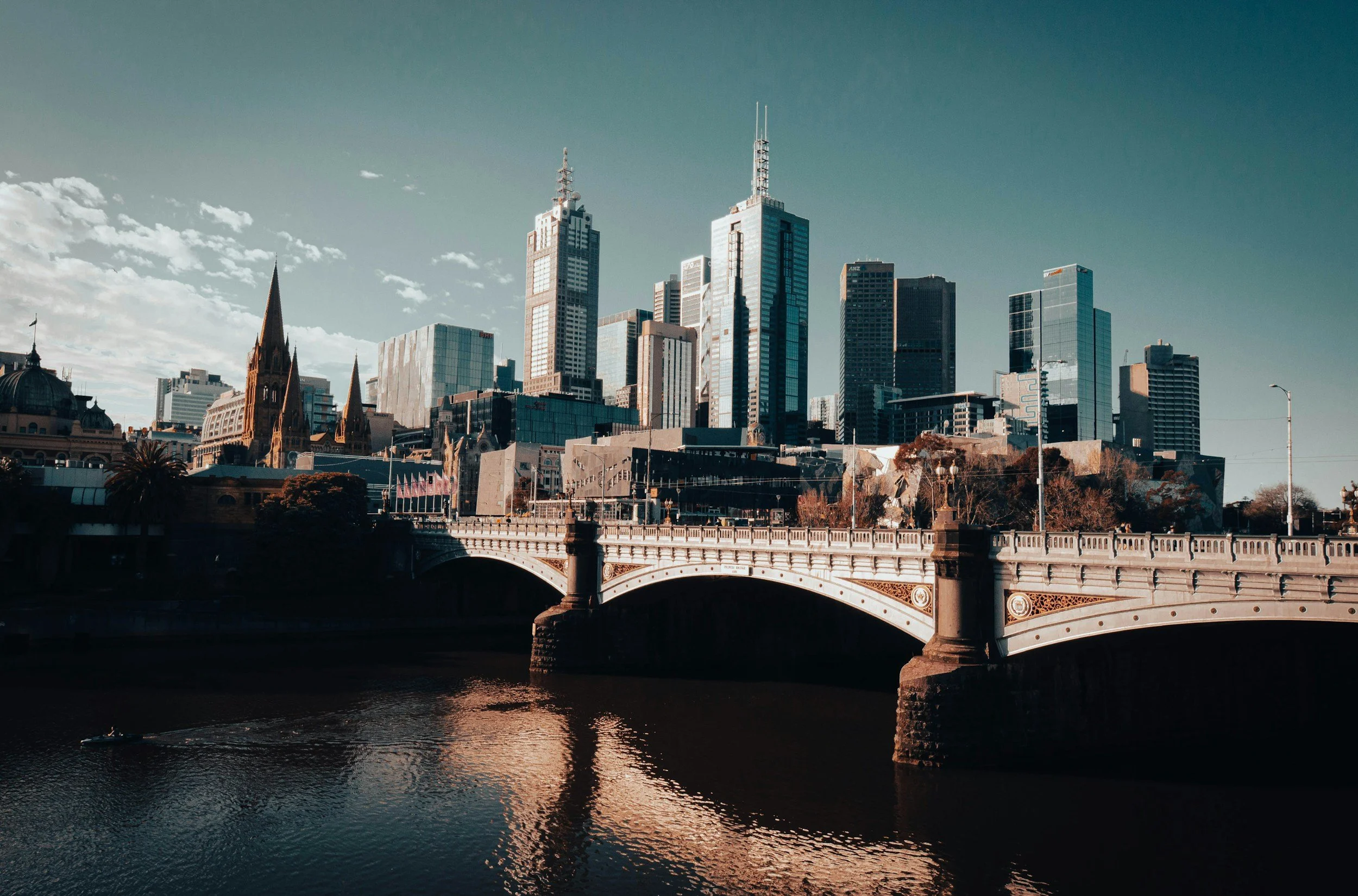 City skyline with modern skyscrapers and historic buildings viewed from a bridge over a river, with a boat on the river.