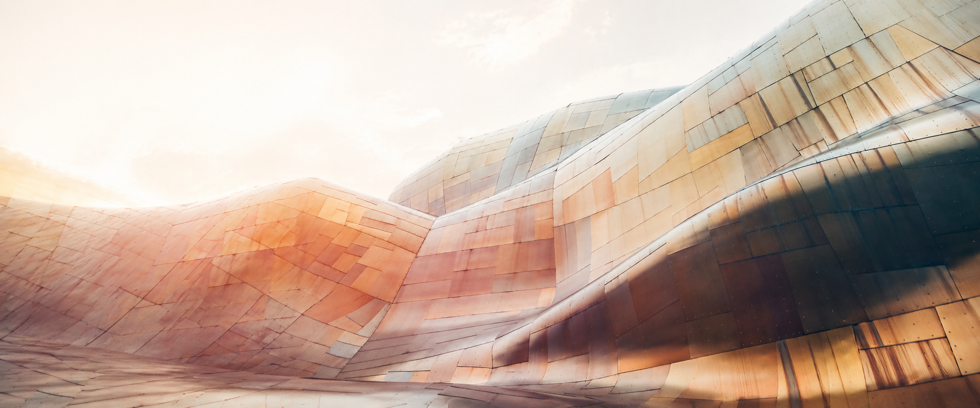 Modern architectural building with flowing, curvilinear design made of metallic panels in shades of copper, bronze, and silver, illuminated by sunlight.
