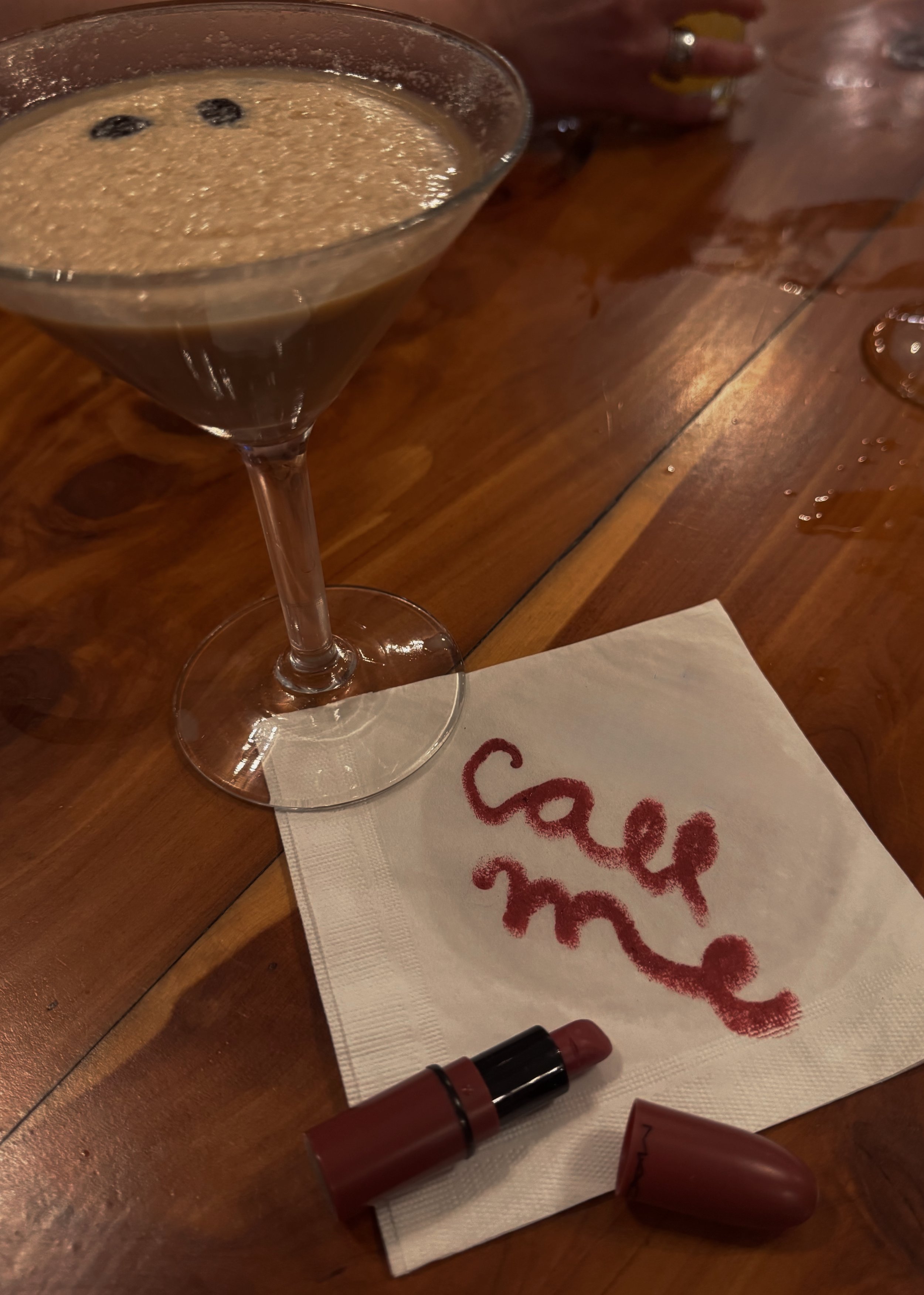 Close-up of a martini glass filled with a brown cocktail, garnished with three dark cherries, resting on a wooden table. In the background, a hand with yellow nail polish and a phone are partially visible. On the table, there is a white napkin with red writing, a brown lipstick tube, and another brown lipstick or tube.