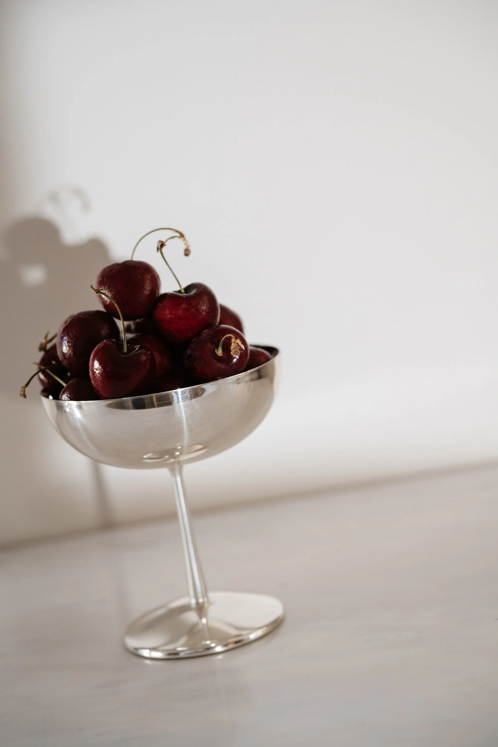 A metallic cocktail glass filled with red cherries, positioned on a white surface against a plain white background.