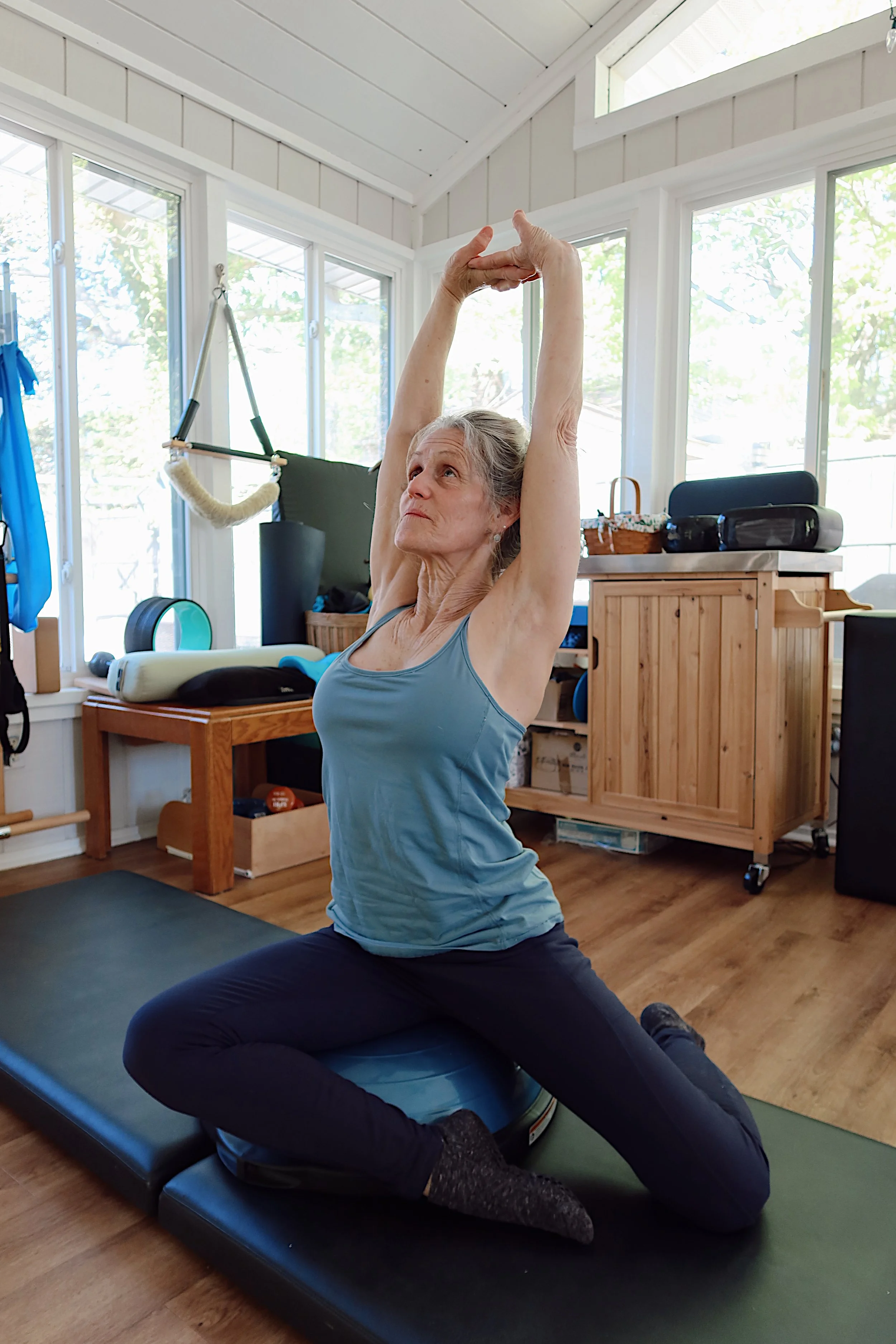 Older woman practicing yoga or stretching indoors on a black yoga mat, with her arms raised above her head and hands clasped. She is wearing a gray tank top and black pants, sitting on her heels with one knee bent. The room has large windows, wooden flooring, and various fitness equipment and furniture visible in the background.