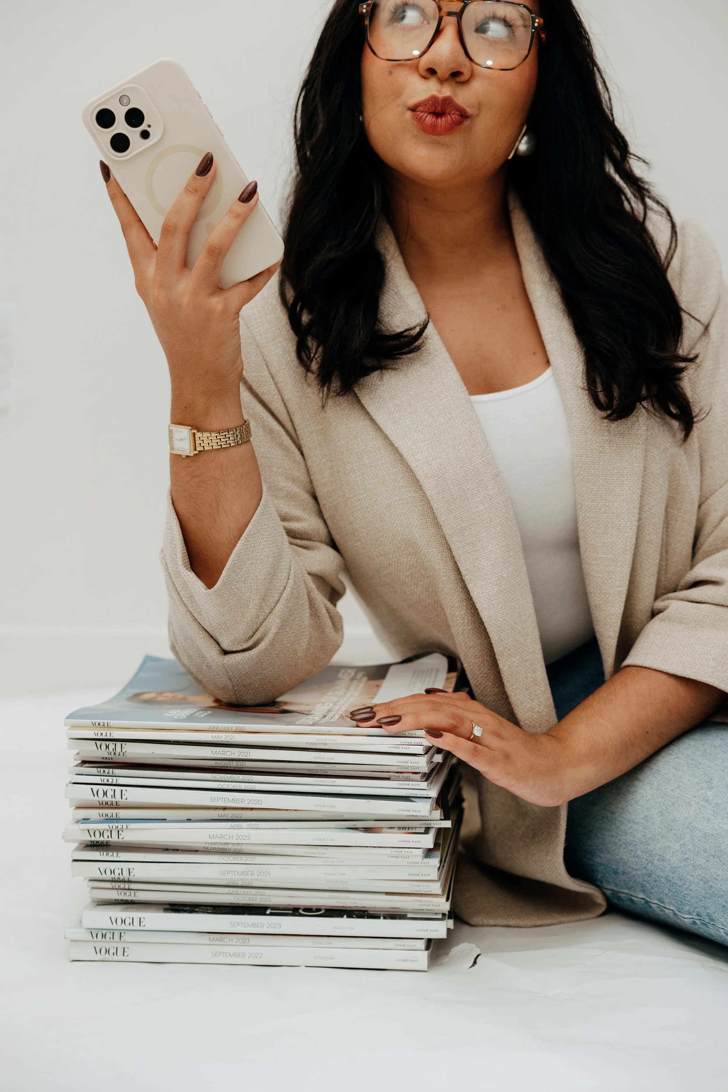 A woman with dark hair, wearing glasses and a beige blazer, holding an iPhone with a white case in her right hand. She is sitting at a table with a large stack of Vogue magazines, and her left hand is resting on the magazines. She is looking up and to the right with her lips puckered.