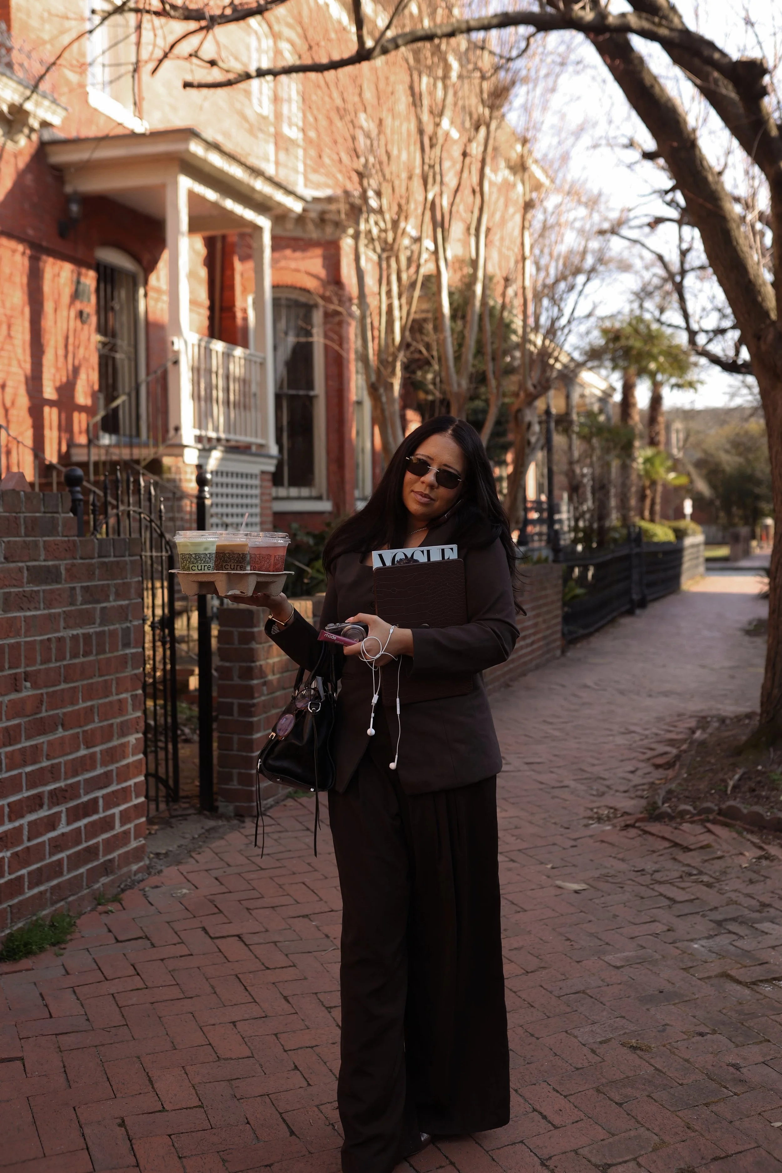 A woman in black outfit and sunglasses is standing on a brick sidewalk, holding a tray with three beverages and a Vogue magazine, in a neighborhood with brick houses, trees, and a lamppost.
