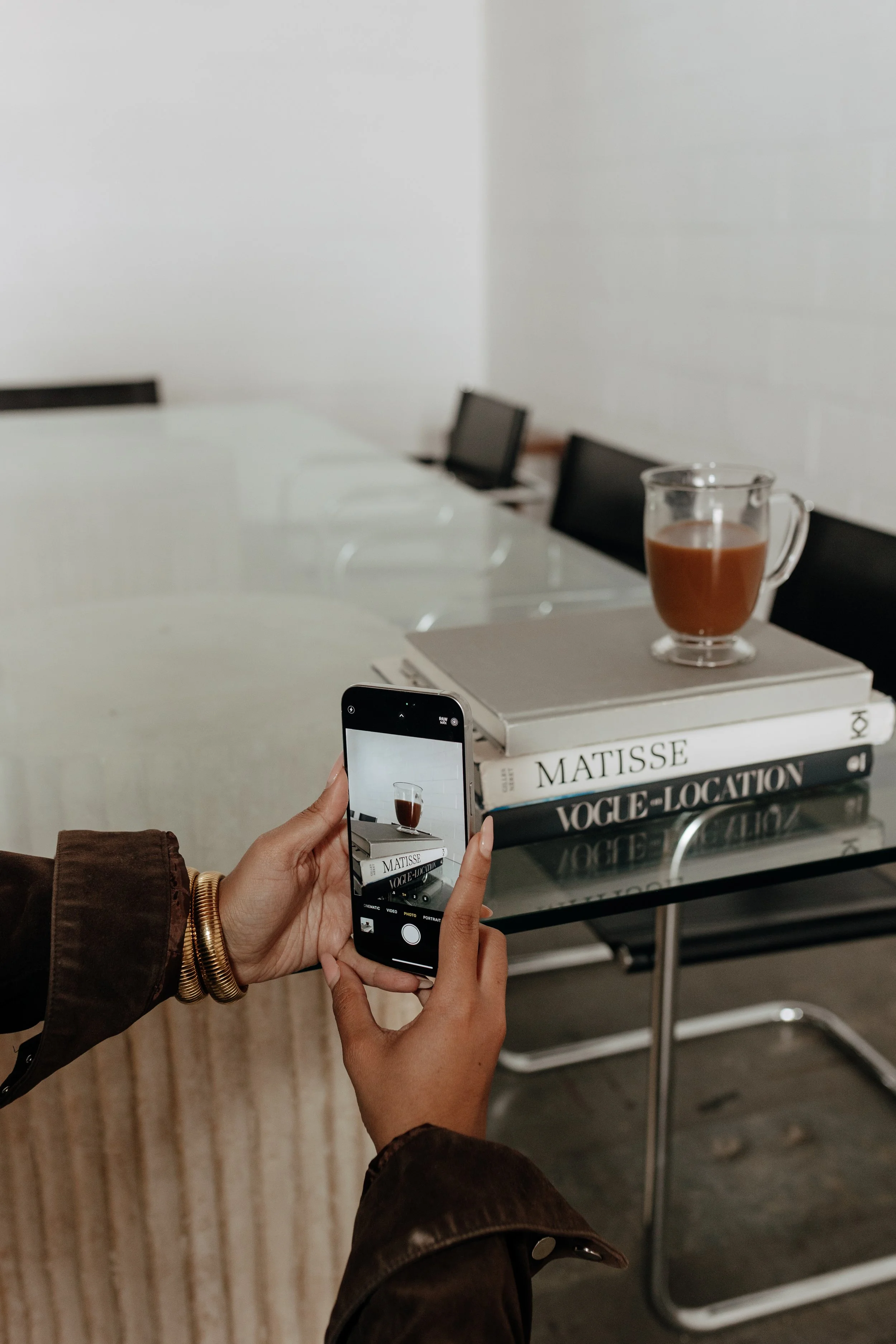 Person taking a photo of a table with a stack of books and a glass of coffee in a modern, minimalistic room.