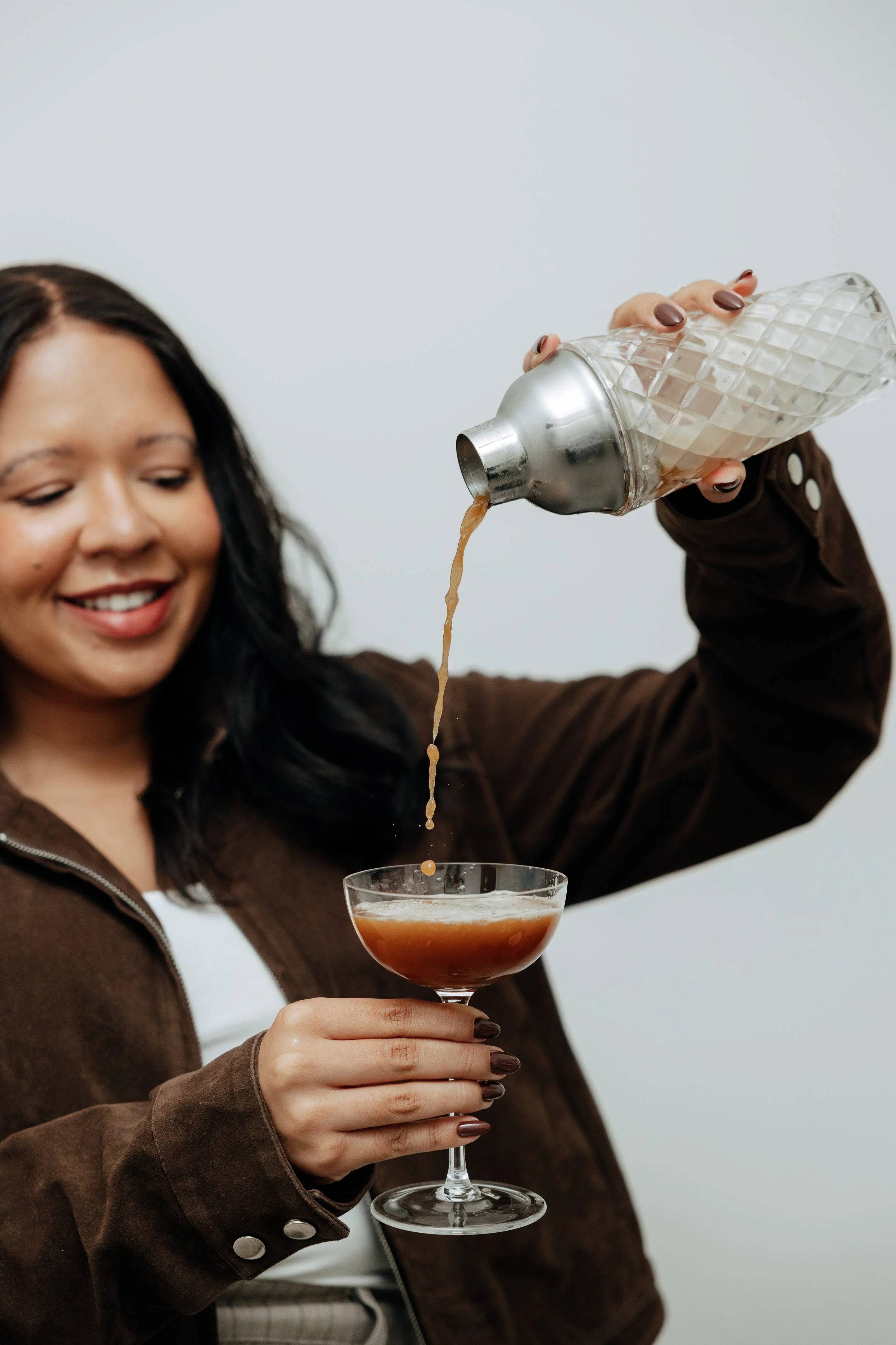 A woman with long dark hair wearing a brown jacket and white shirt is pouring a cocktail from a textured glass shaker into a coupe glass, holding it with her left hand. She is smiling with her eyes closed.