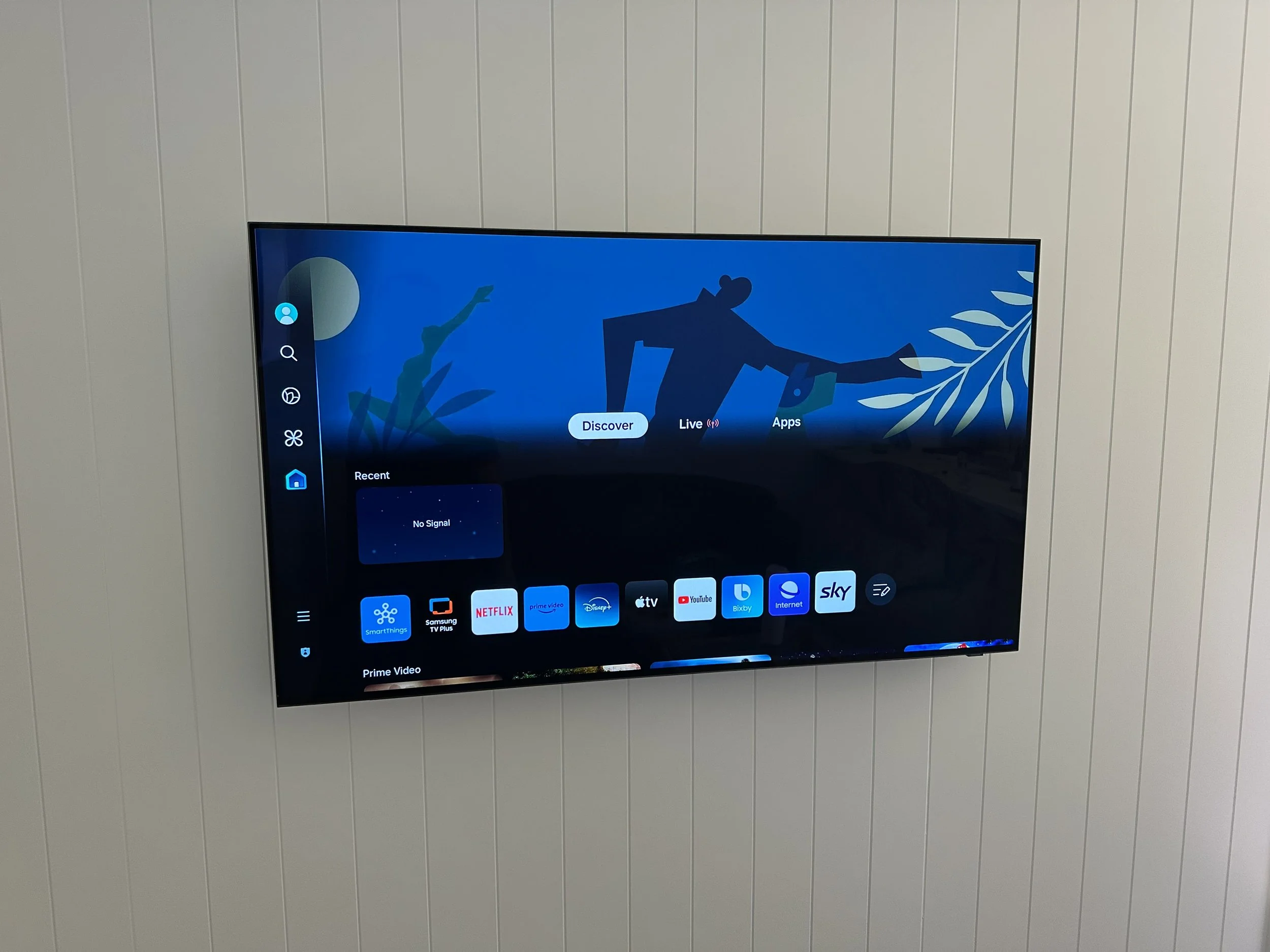 Flat-screen television mounted on a wall displaying a menu with streaming apps such as Netflix, Prime Video, Disney+, YouTube, and others, on a dark background with blue and green artwork.