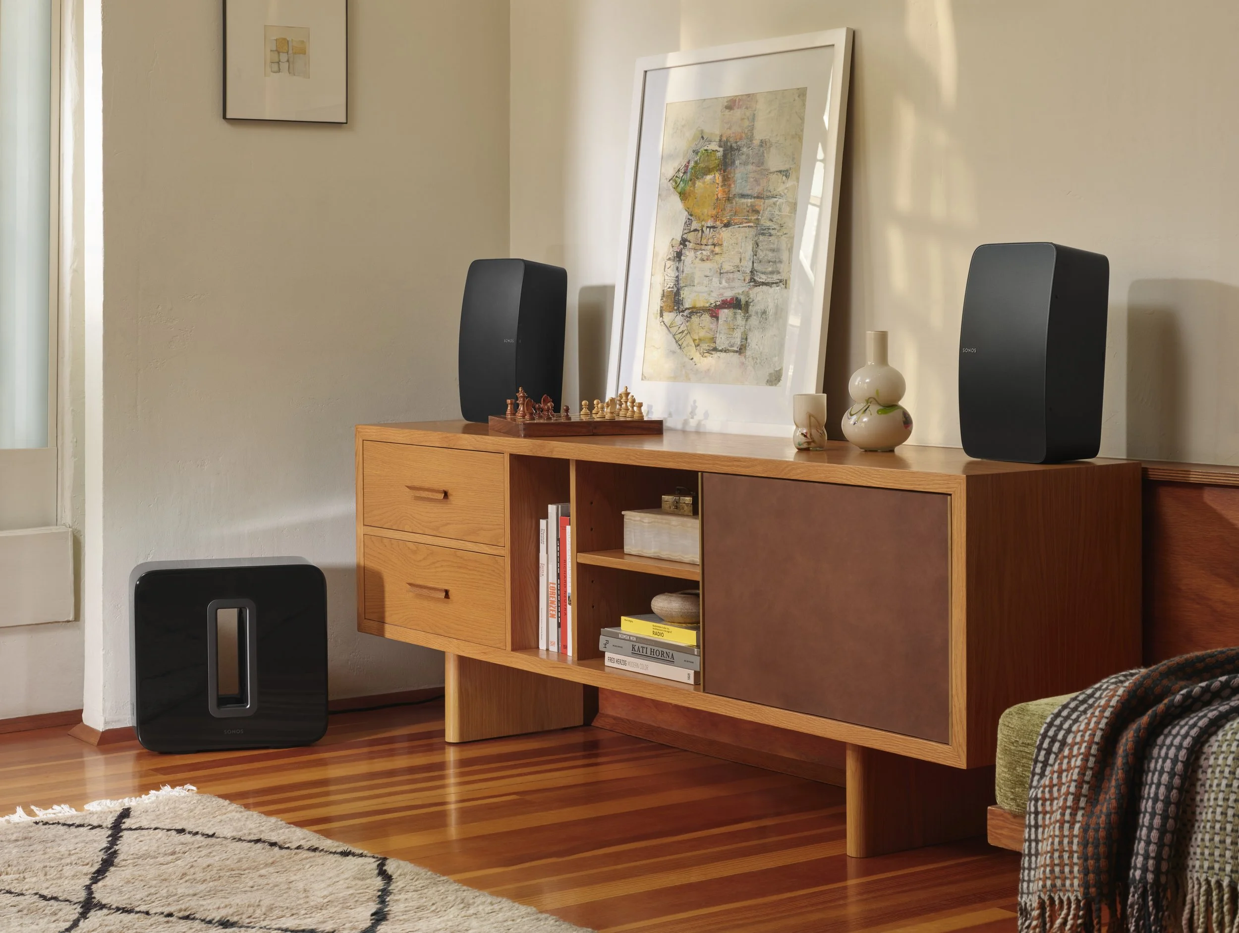 Living room corner with a wooden media console holding two black speakers, decorative vases, and a framed art print, with a rug on polished hardwood floor.