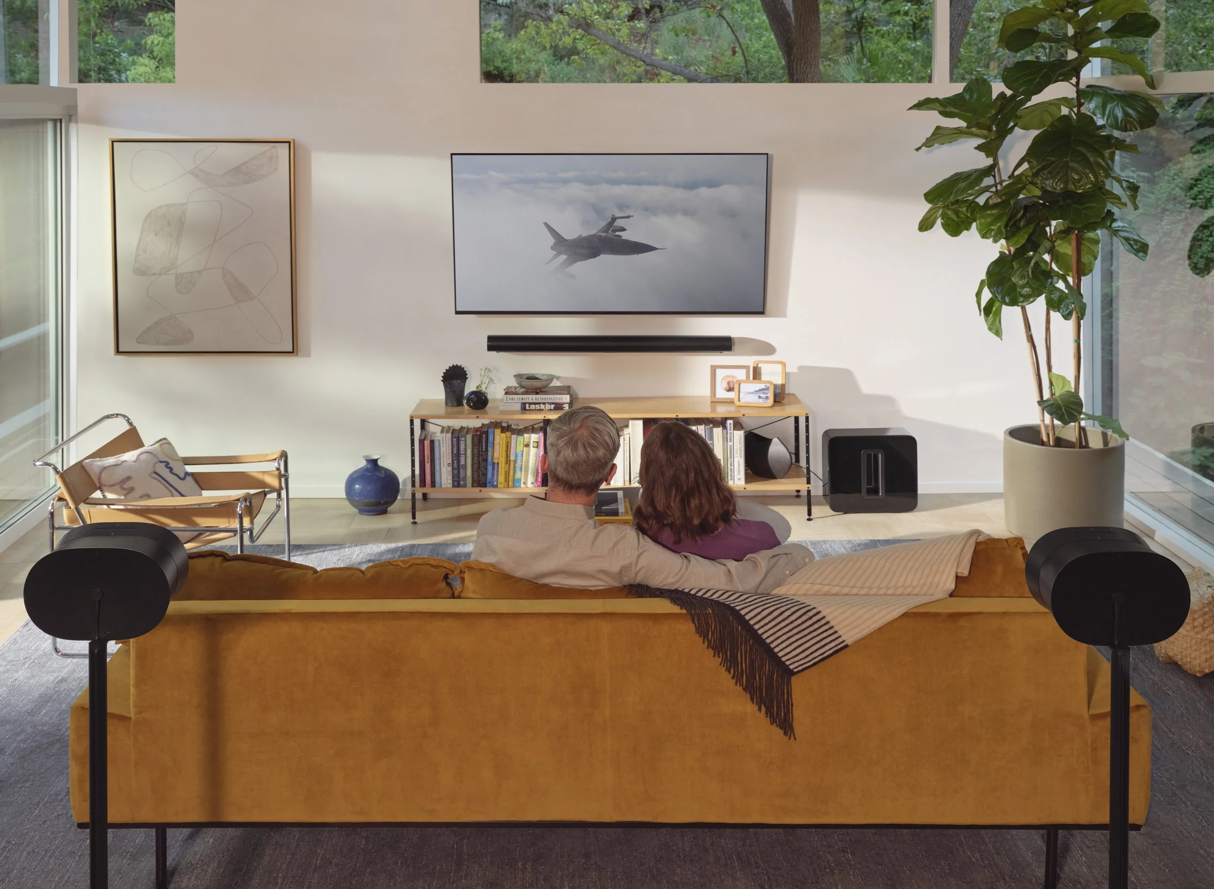 A couple sitting on a mustard yellow sofa in a modern living room, watching a flat-screen TV showing a fighter jet in the sky. The room has large windows, a tall potted plant, and various decorative items, including books, framed photos, and artwork.