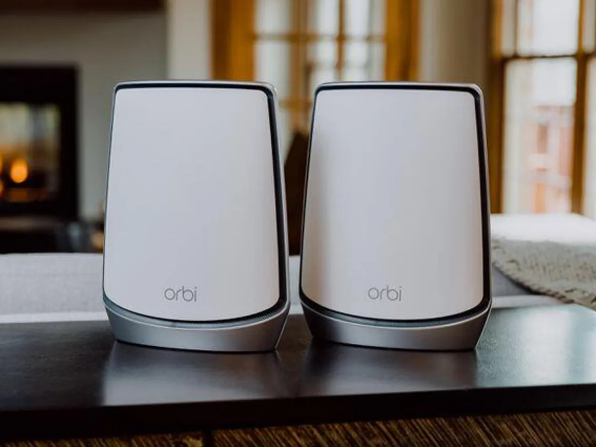Pair of white Orbi Wi-Fi speakers on a dark surface in a cozy living room with warm natural lighting.