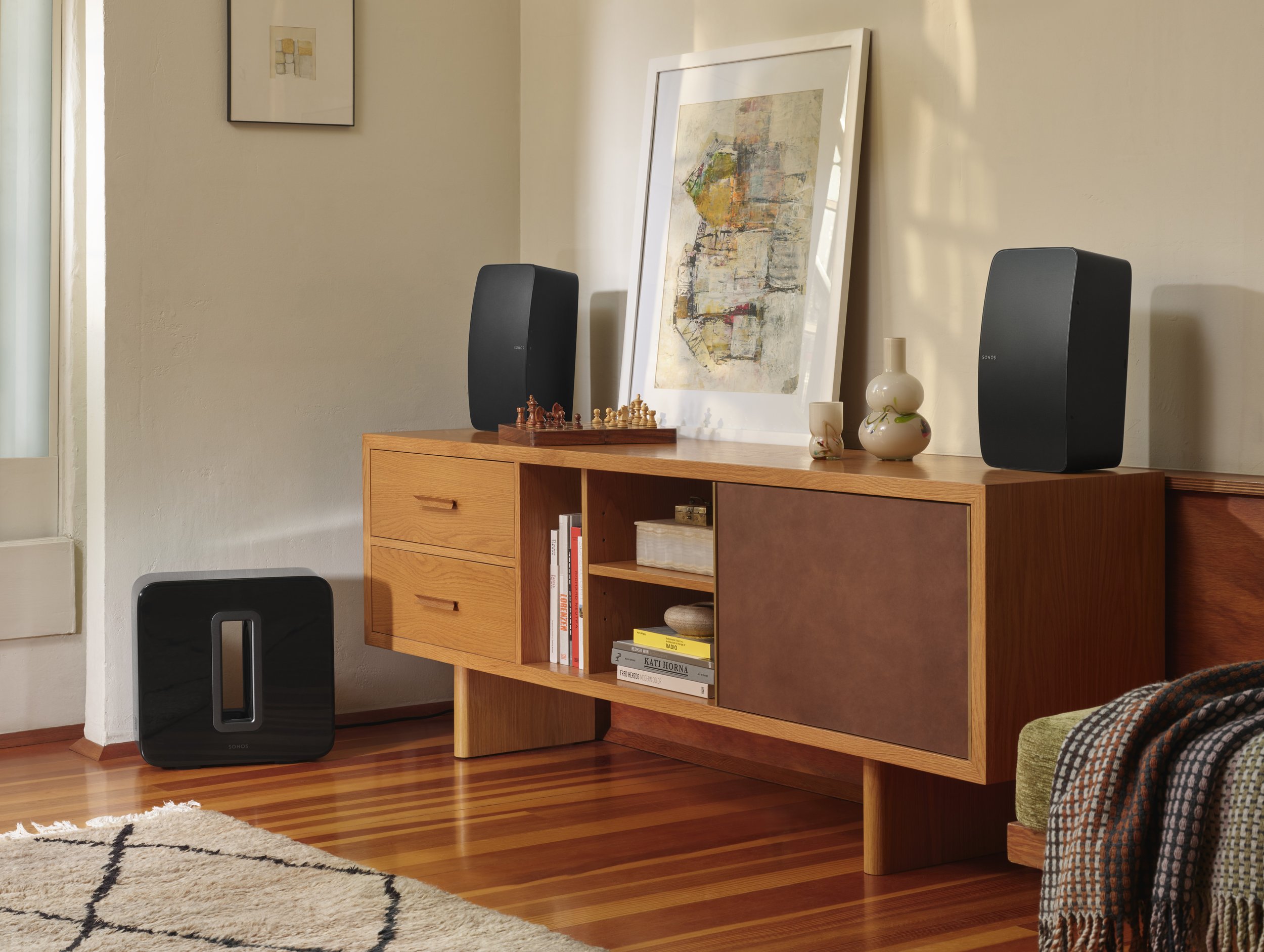 Living room sideboard with black Sonos speakers, decorative vases, a framed abstract art piece, books, and a chess set, with a window, a rug, and a green armchair nearby.