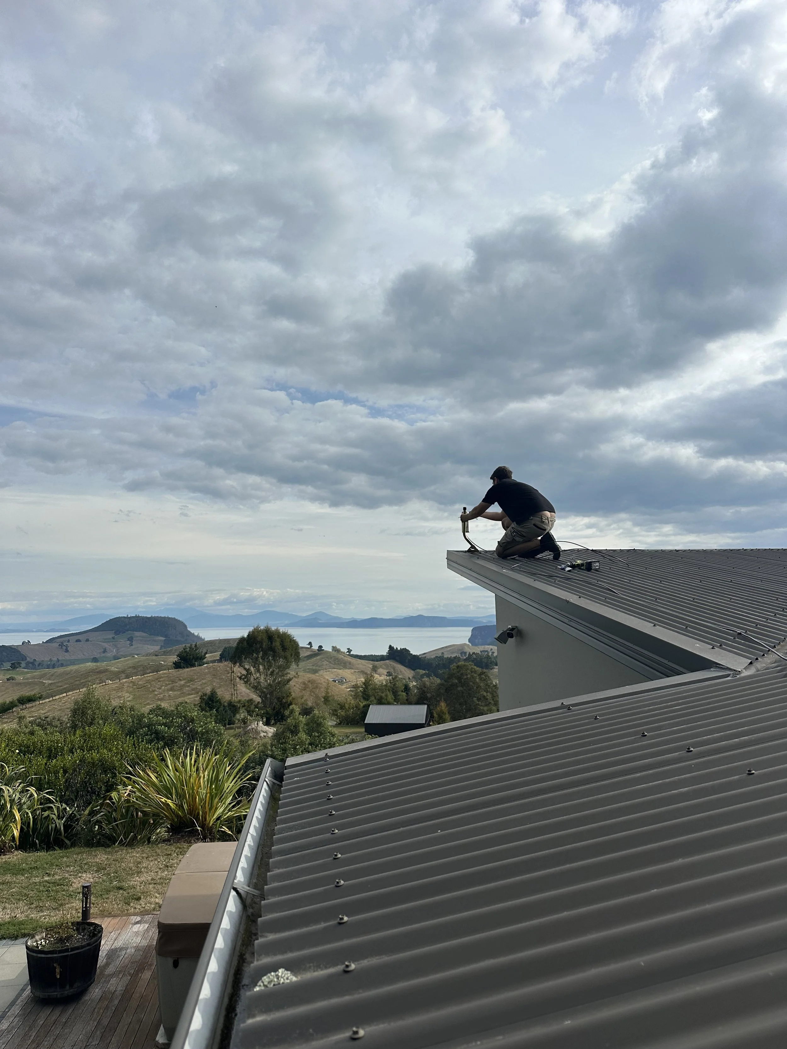 A man is kneeling on a roof, using a tool, with a scenic landscape of hills, trees, and water in the background under a cloudy sky.