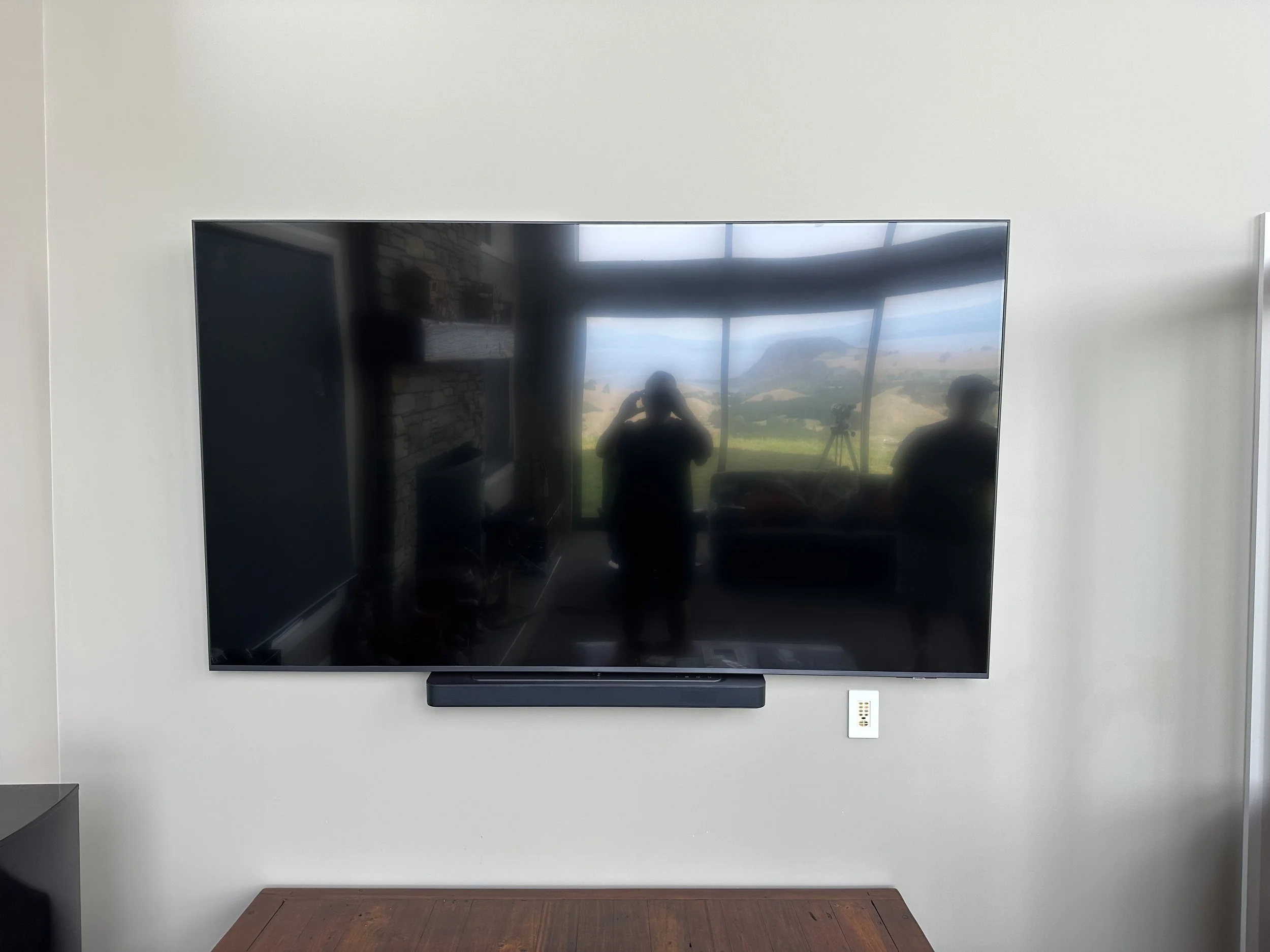 Wall-mounted flat-screen TV with reflection of interior and outdoor view, soundbar underneath.