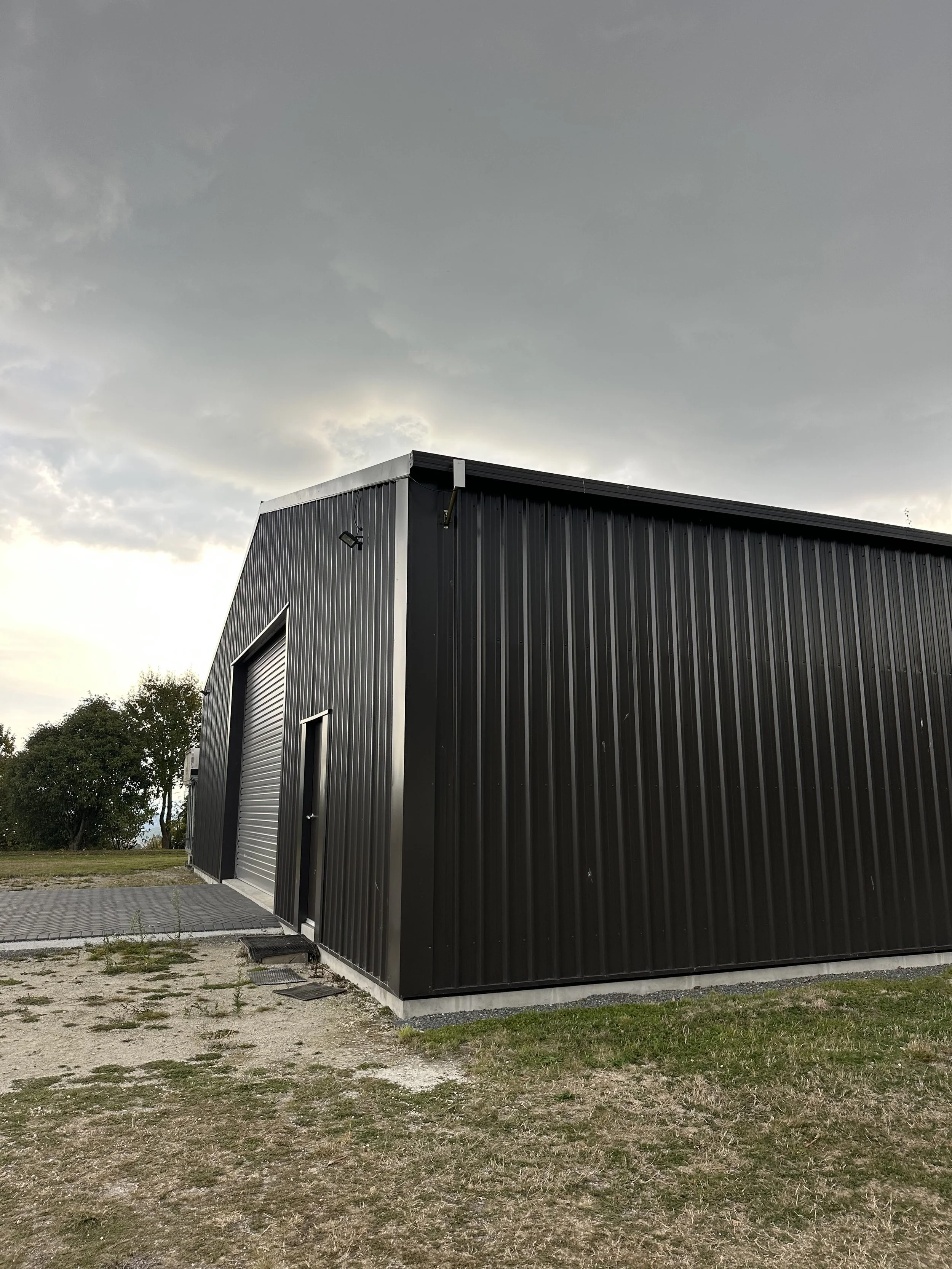 Large gray metal building with a roll-up door and small entry door against a cloudy sky, surrounded by grass and paving stones.