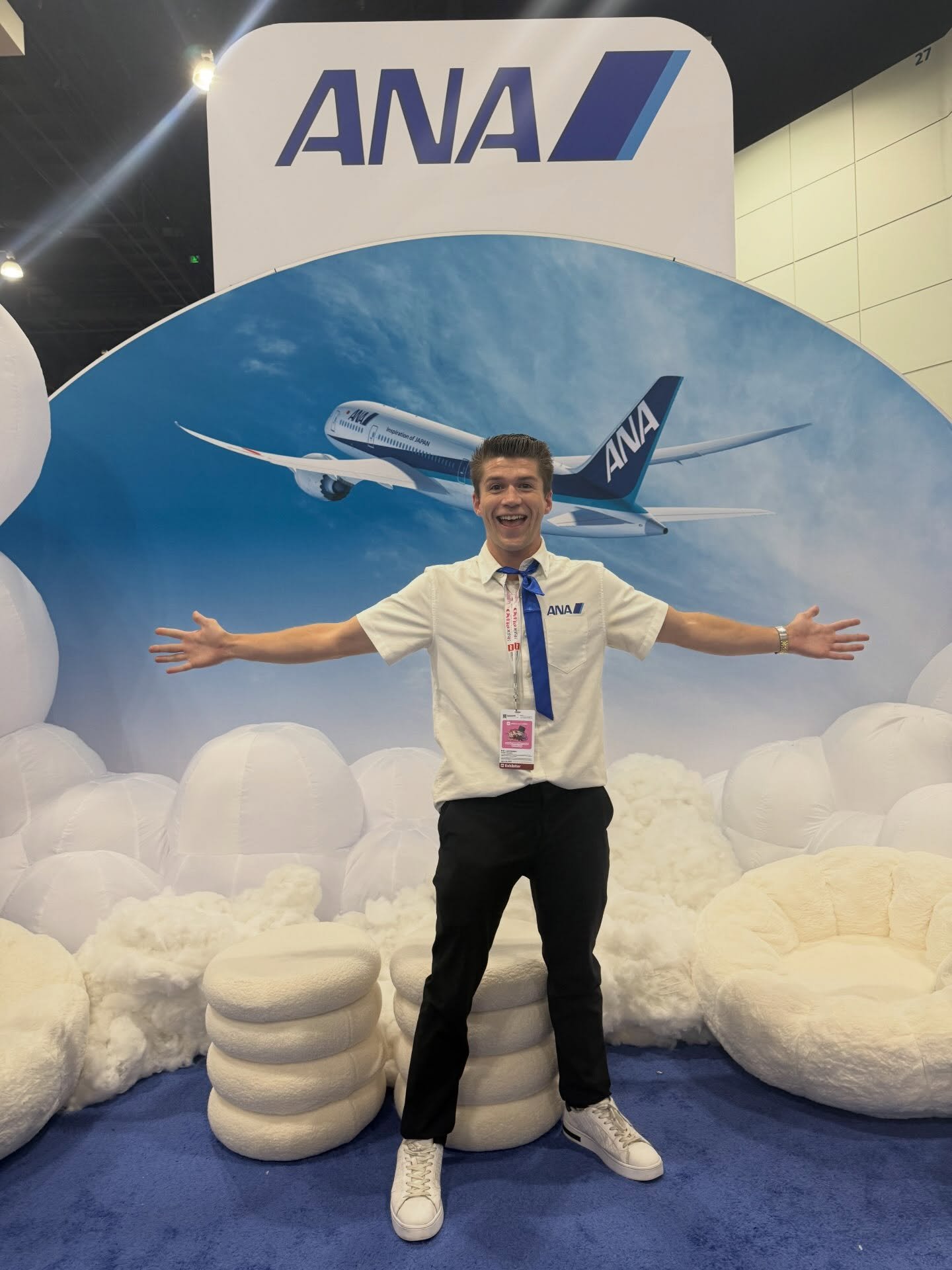 A man with a short hairstyle, wearing a uniform with the ANA logo, smiling with arms outstretched, standing in front of a large ANA airplane poster surrounded by inflatable cloud-like structures.