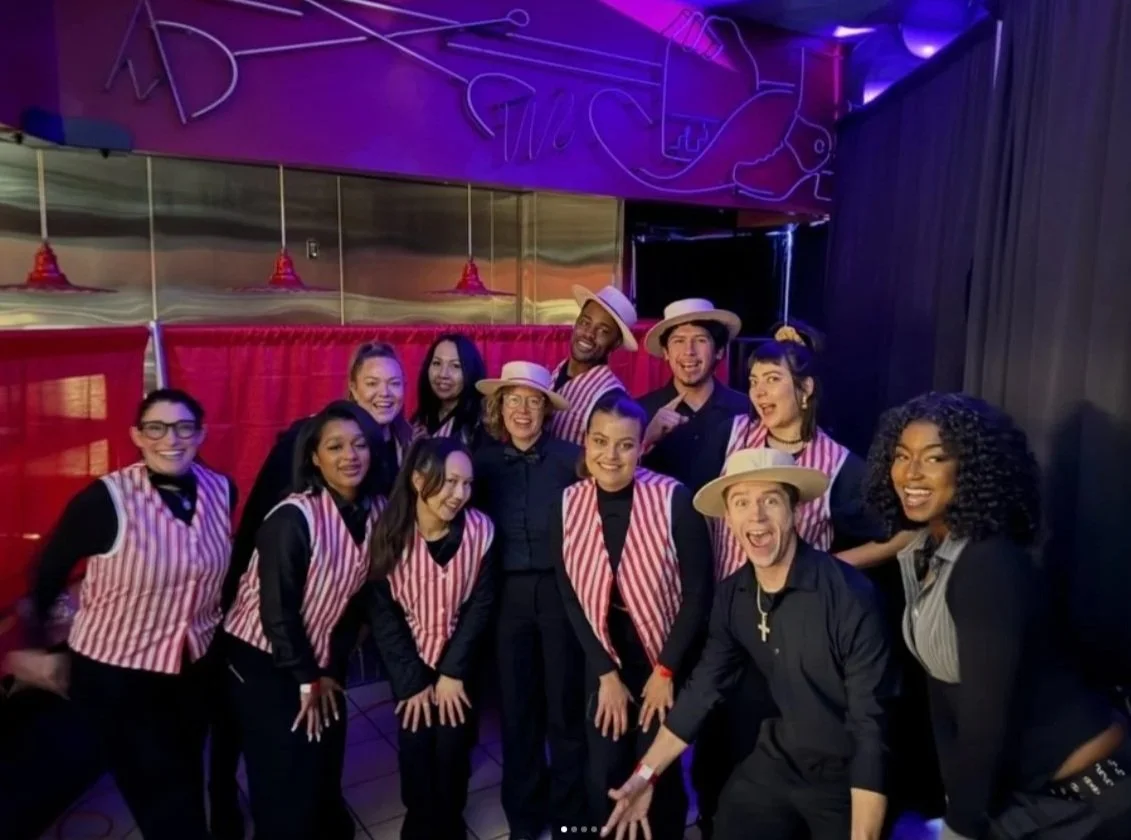 Group of people dressed as waitstaff, some wearing striped aprons and hats, posing together in a restaurant or bar with neon lights and red curtains in the background.