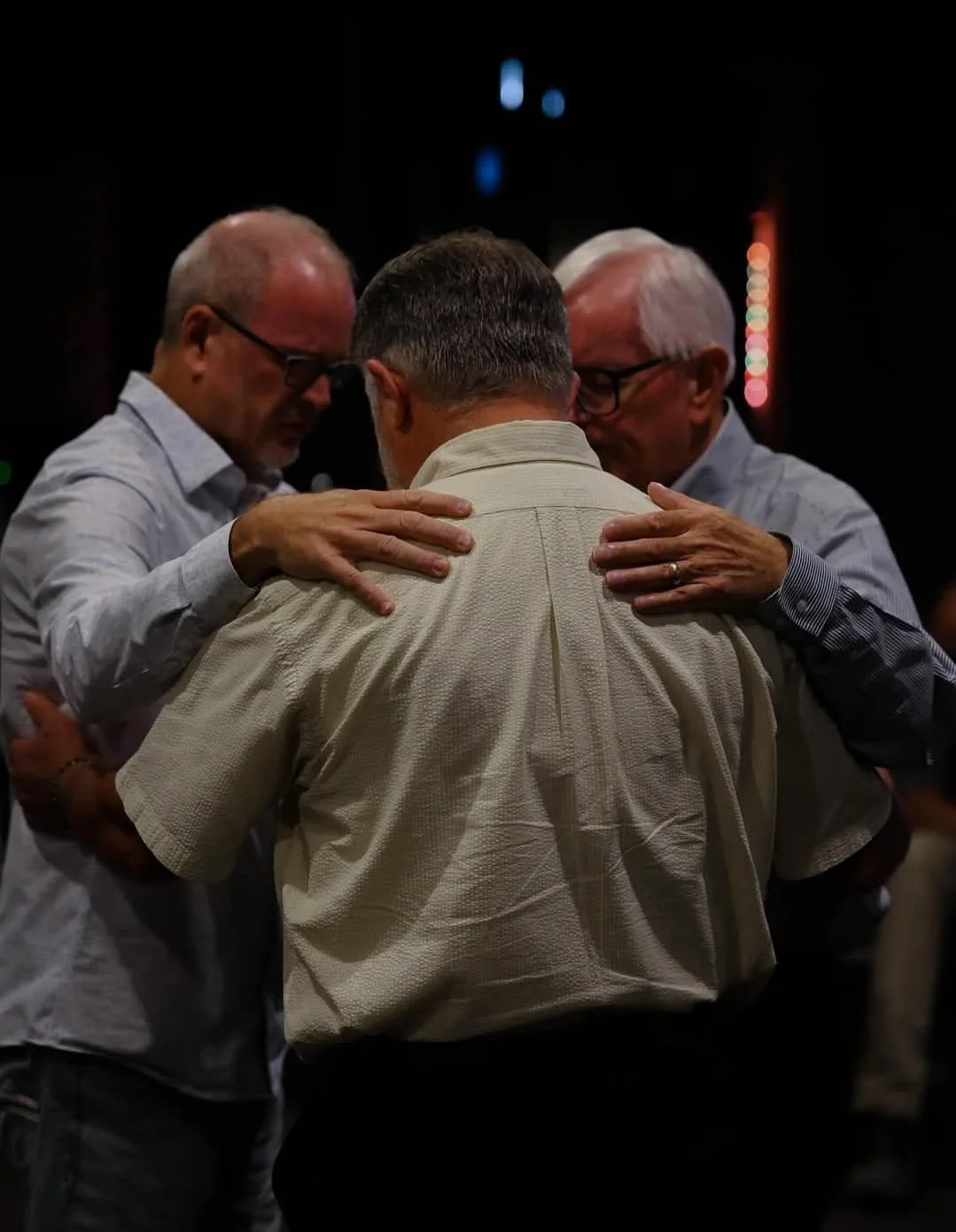 Three men praying or comforting each other, huddled close with their heads bowed.