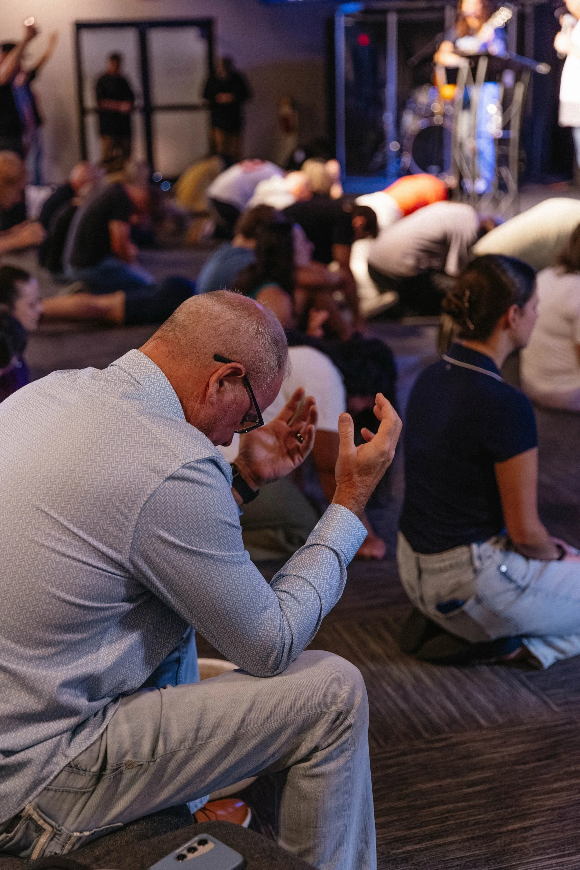 People kneeling and praying during a religious gathering or worship, with some individuals with raised arms, set indoors with a small stage and musical equipment in the background.