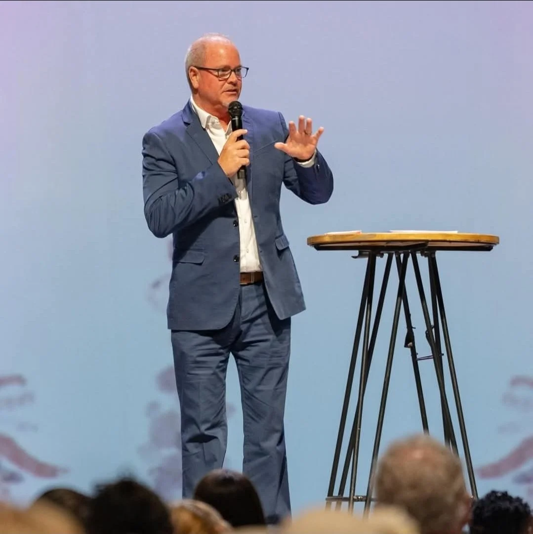 A man with glasses and short gray hair, dressed in a blue suit and white shirt, stands on stage holding a microphone and gesturing with his left hand. There is a small round table to his right, and the background is a plain light blue wall, with audience members visible at the bottom of the image.
