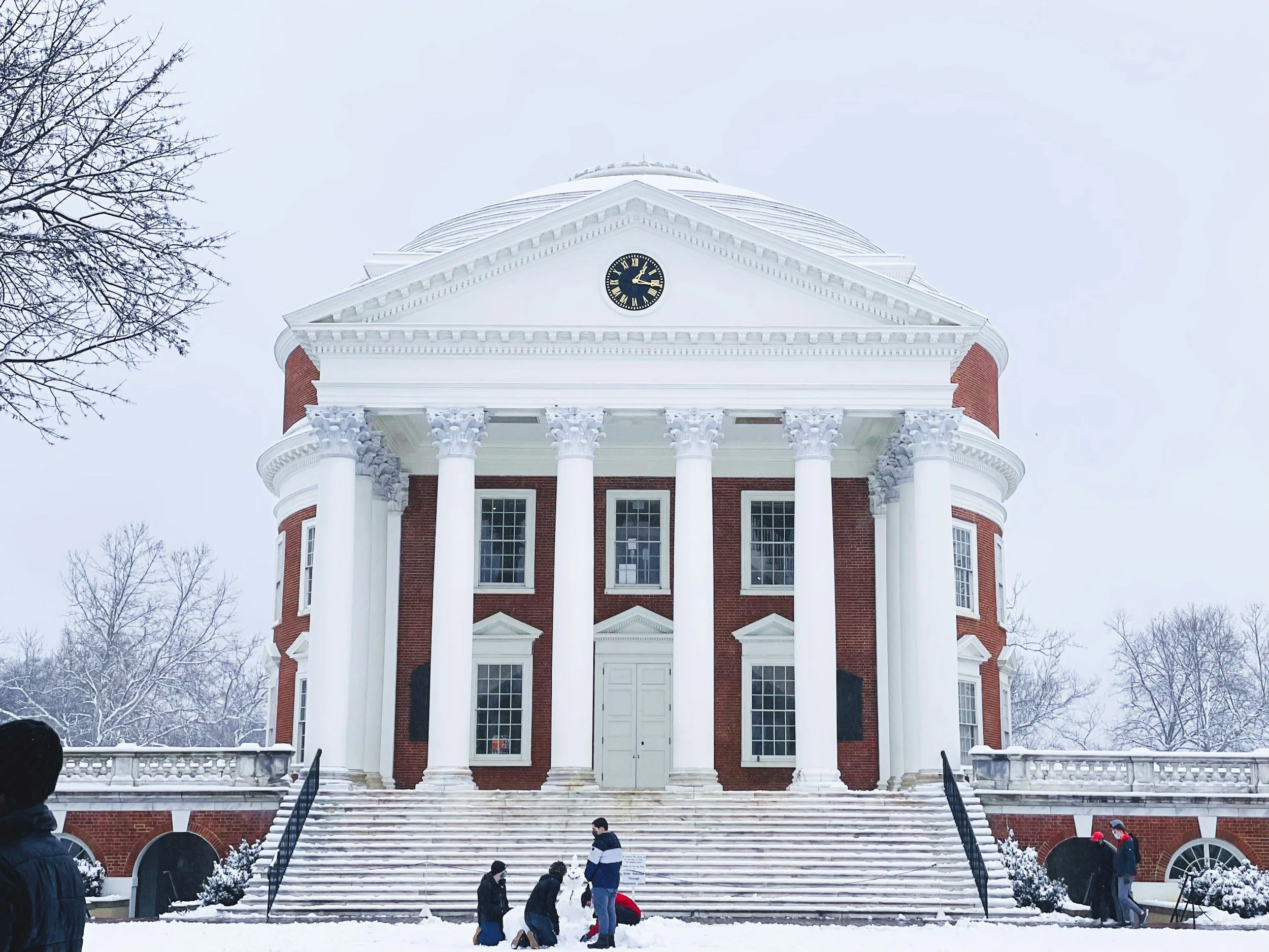 A red brick building with white columns, a clock showing 4:17, and white stairs leading up to it, set in a snowy landscape with leafless trees and people dressed in winter clothing.