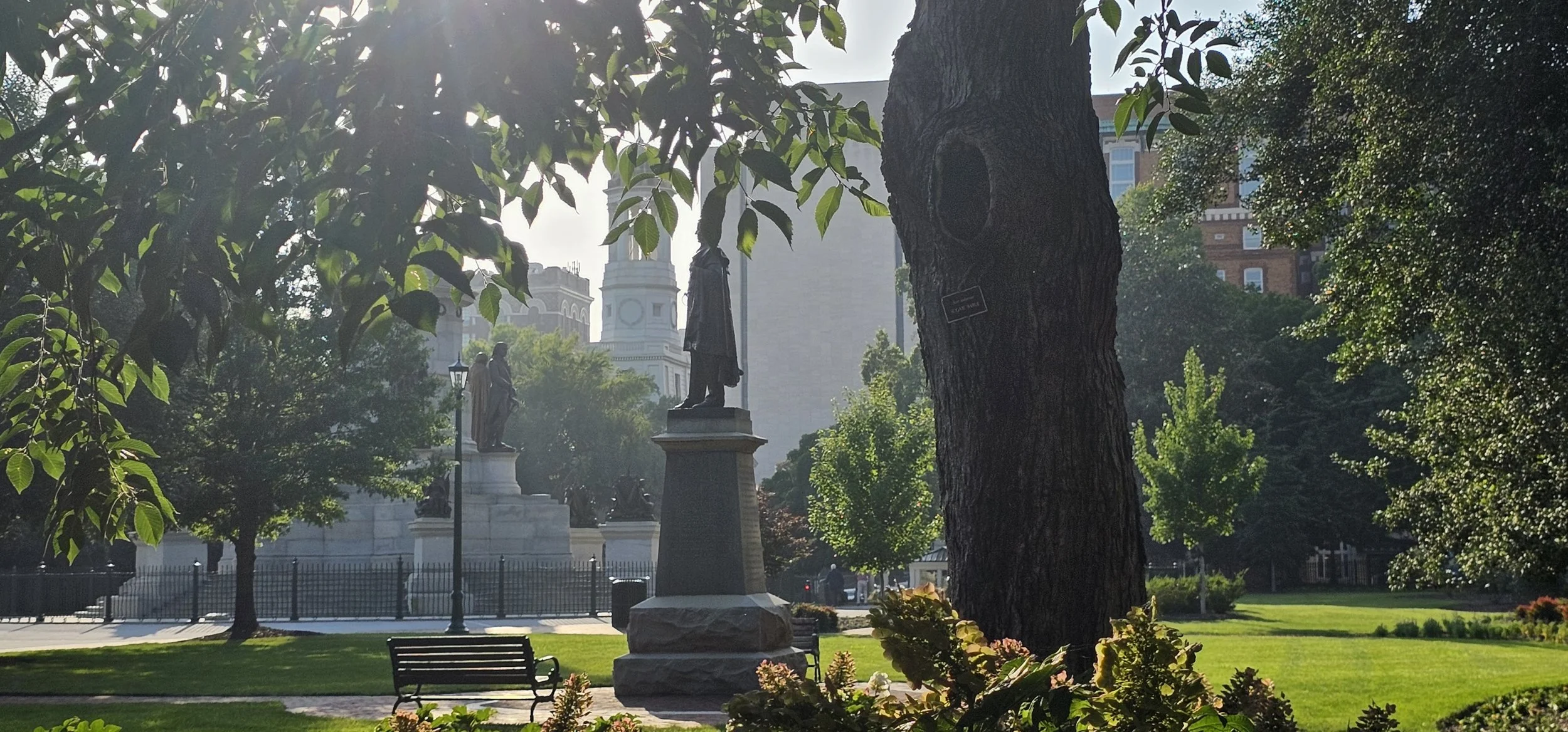 A park with statues, trees, benches, and a city skyline in the background, with the sun shining through the leaves.