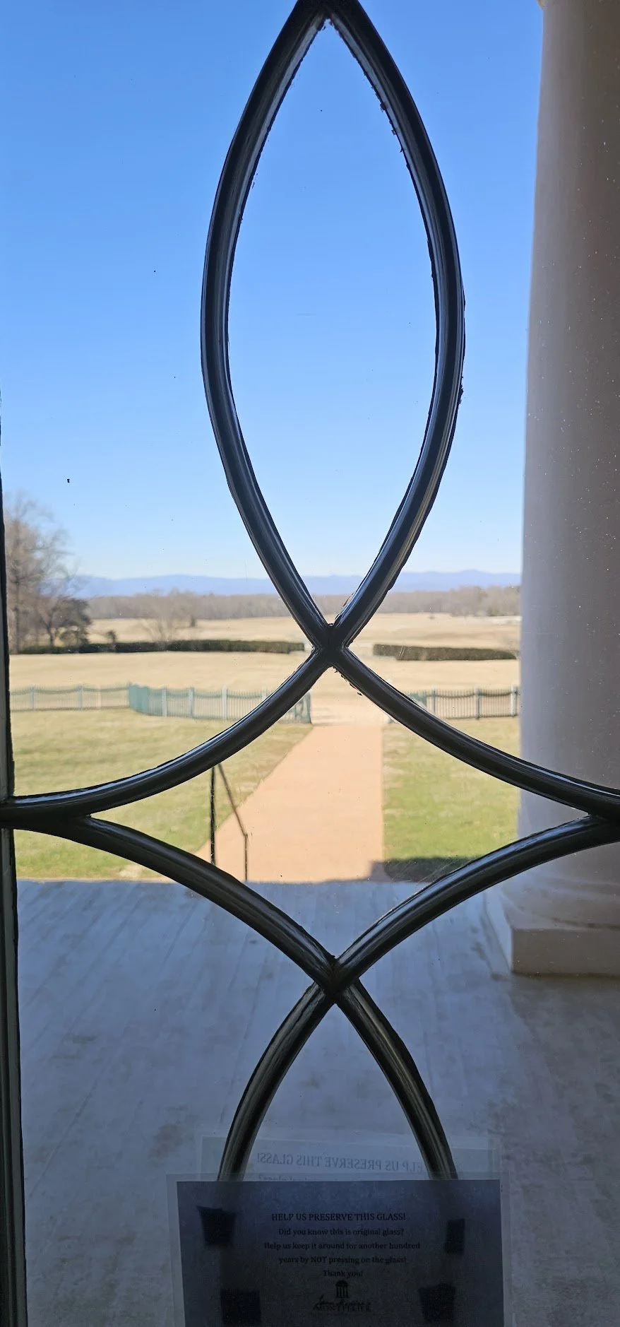 View of a grassy field and distant hills through a window with decorative metal grilles shaped like loops, with a sign on the lower part of the glass.