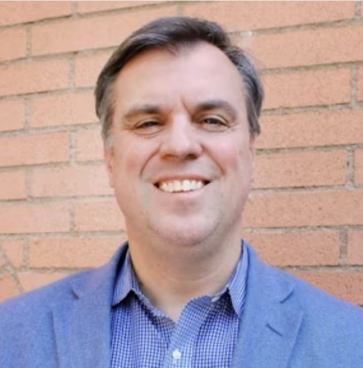 A smiling man standing in front of a brick wall, wearing a blue blazer and a blue checkered dress shirt.