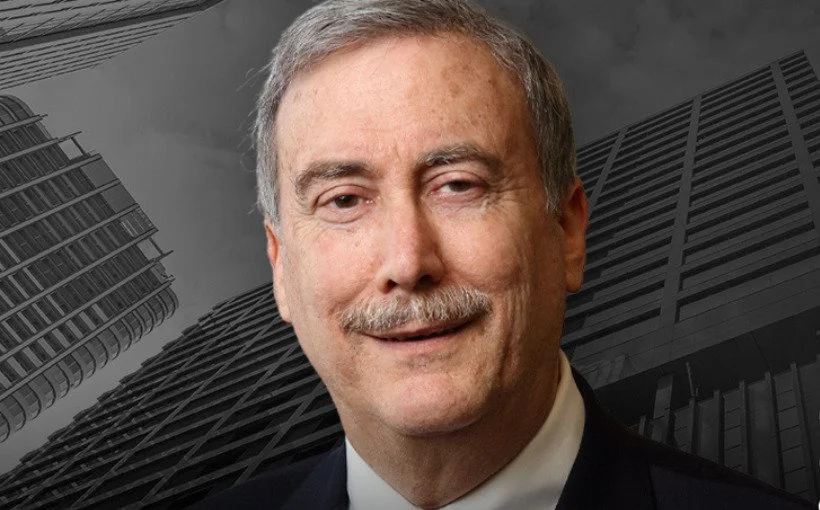 Portrait of Larry Sabato with gray hair, a mustache, and a slight smile, wearing a suit and tie, against a backdrop of modern skyscrapers.