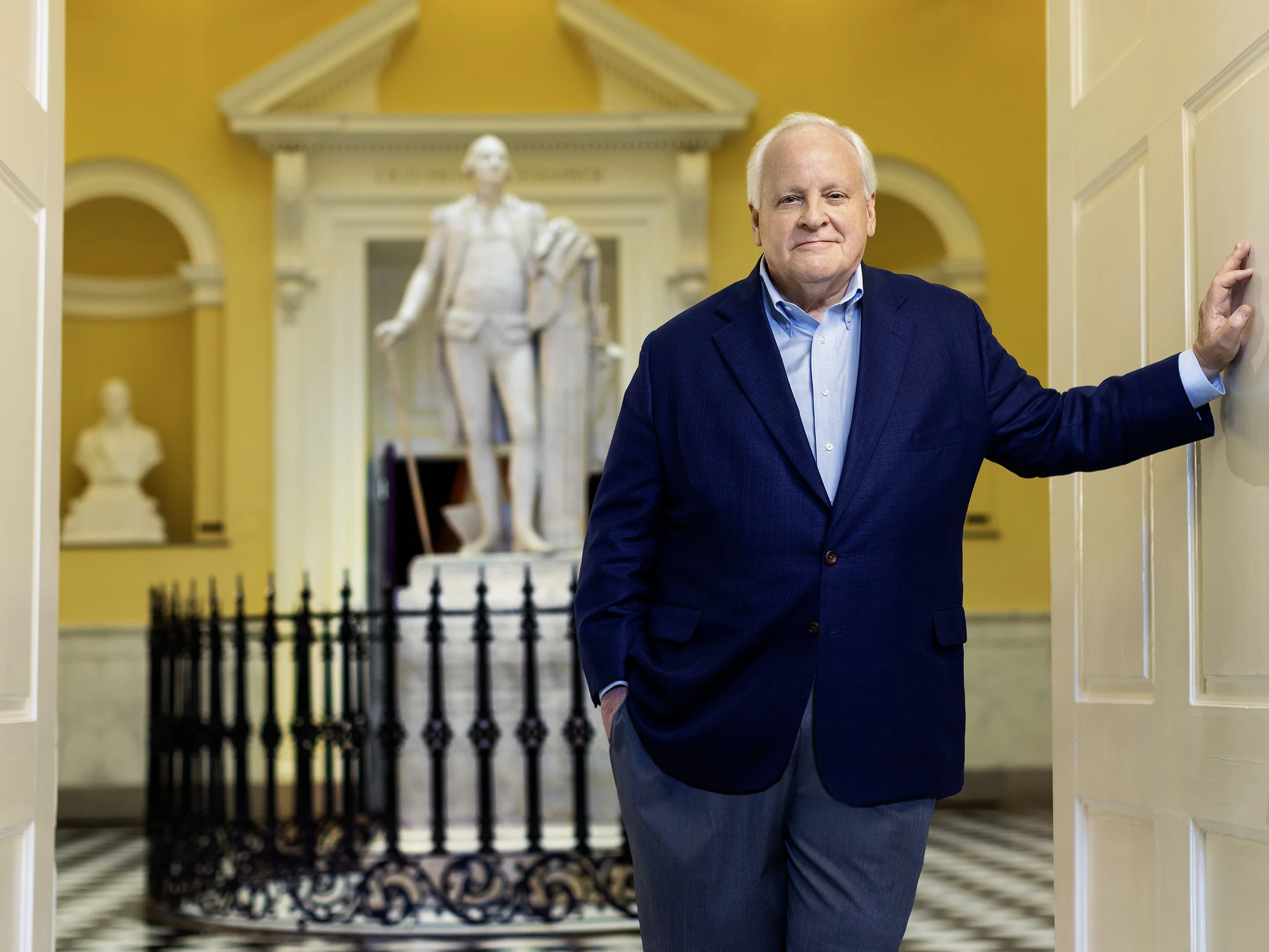An older man in a blue suit is standing in doorway with his hand on the wall, in a room with yellow walls and historic statues and busts displayed in alcoves, including a large statue of a man in a coat and holding a hat.
