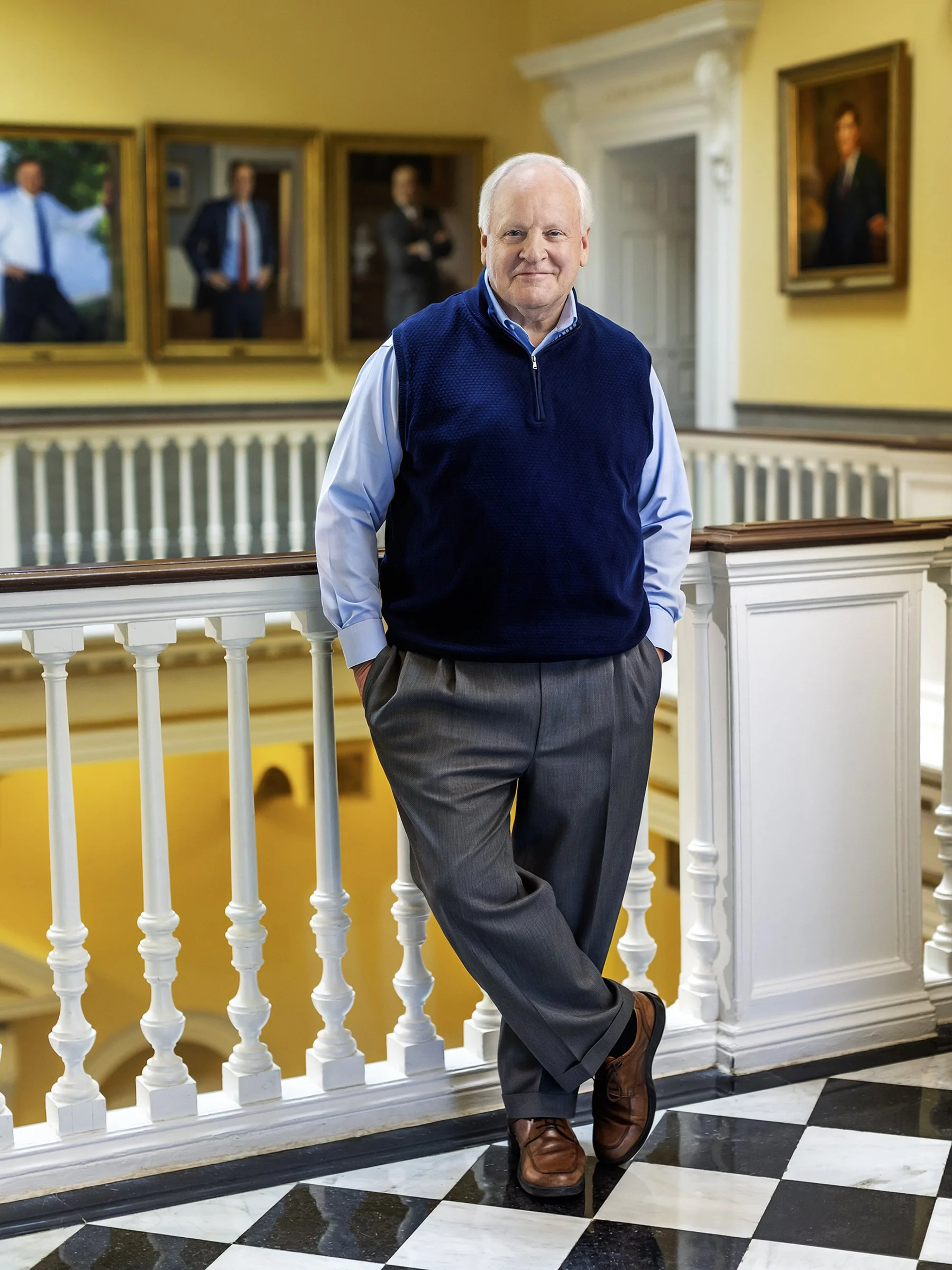 A man with white hair standing on a balcony railing inside a building, dressed in a blue sweater vest, light blue shirt, gray wool pants, and brown shoes, with paintings hanging on the wall behind him and black-and-white checkered marble flooring.