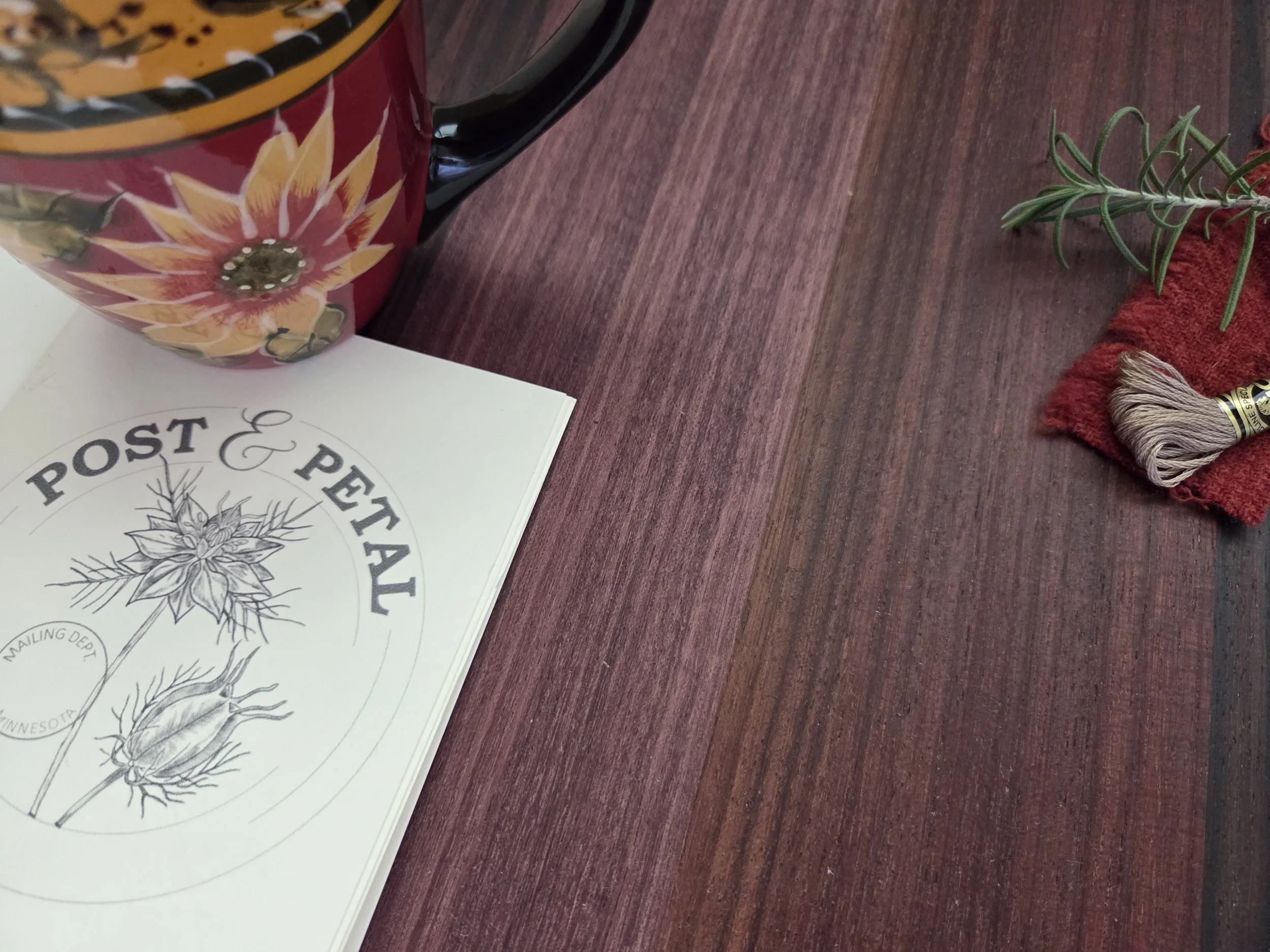 A top-down lifestyle photograph featuring a white card with a botanical logo for 'Post and Petal,' accompanied by a sprig of fresh rosemary, a spool of taupe embroidery thread on a swatch of red wool, and a decorative floral mug in the background.