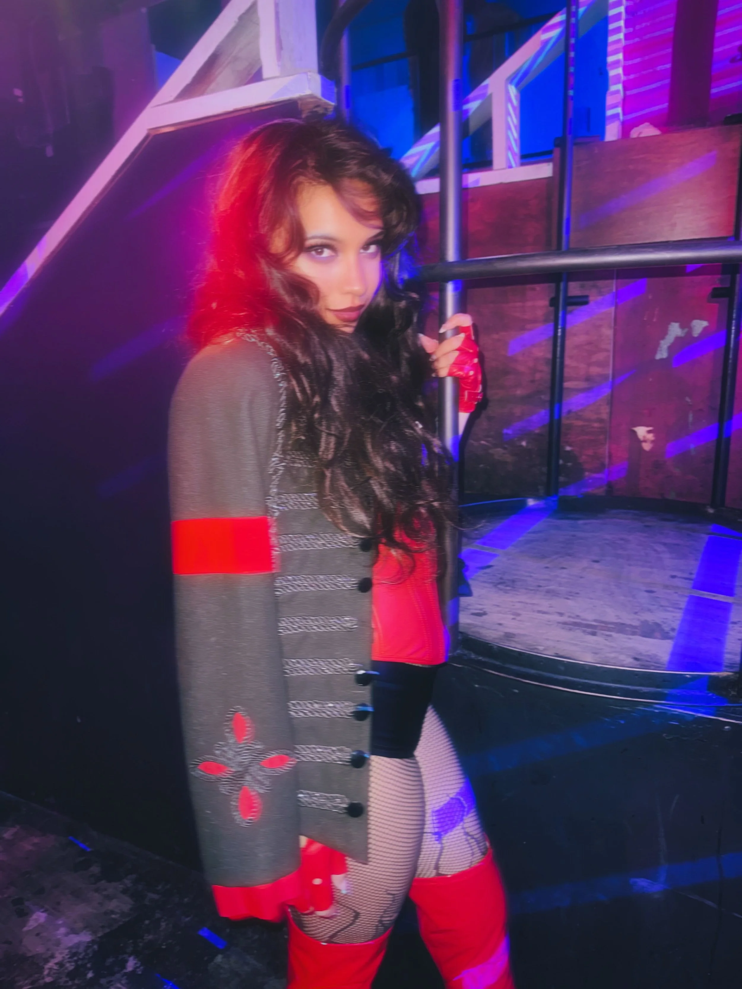 A woman with long dark hair and makeup wearing a red and black outfit with fishnet stockings, standing near a metal railing with colored lighting in the background.