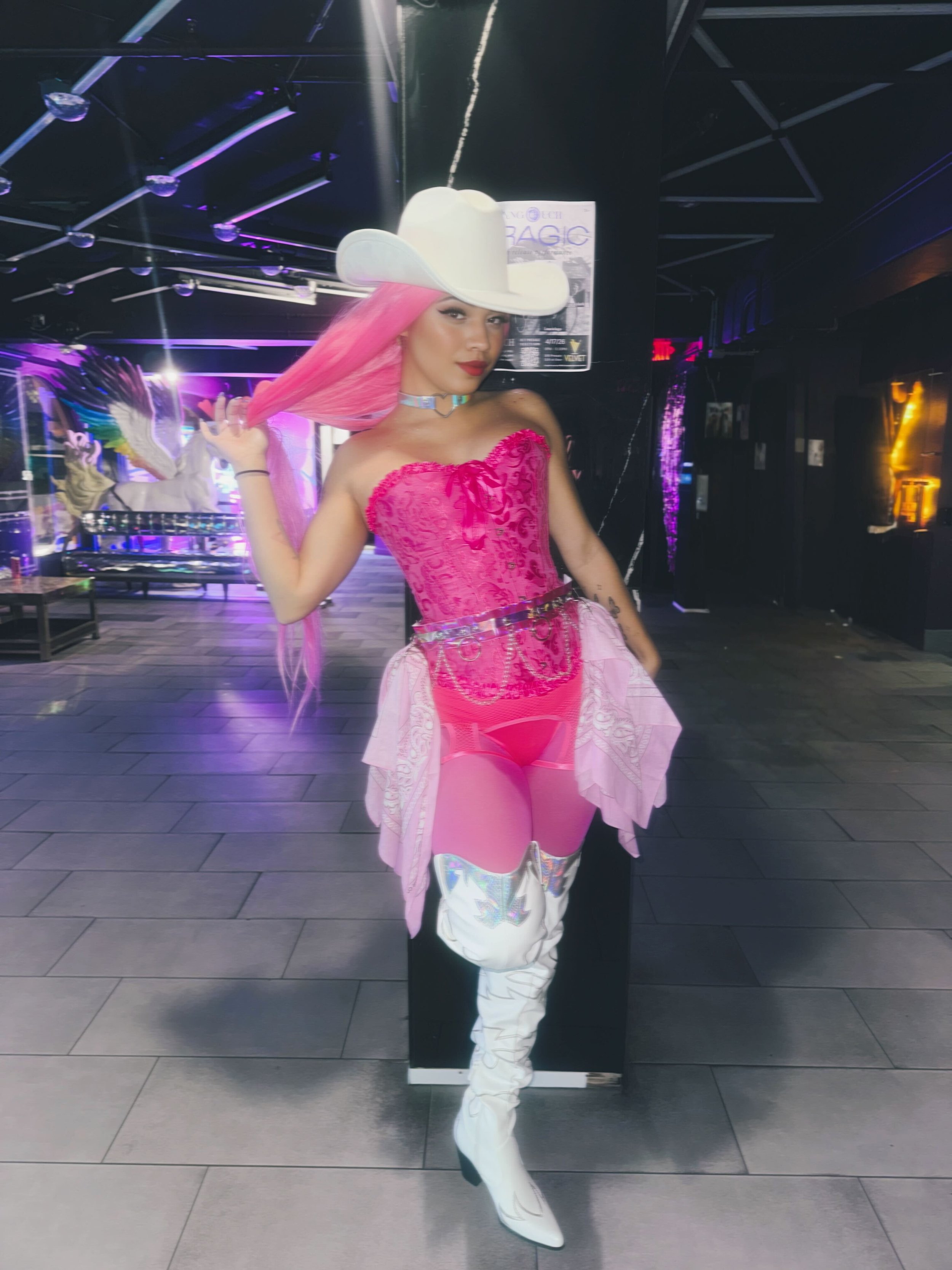 Person with pink hair wearing a large white cowboy hat, pink corset, pink skirt, and white high-heeled boots standing in a dimly lit room with neon lights and a unicorn mural in the background.