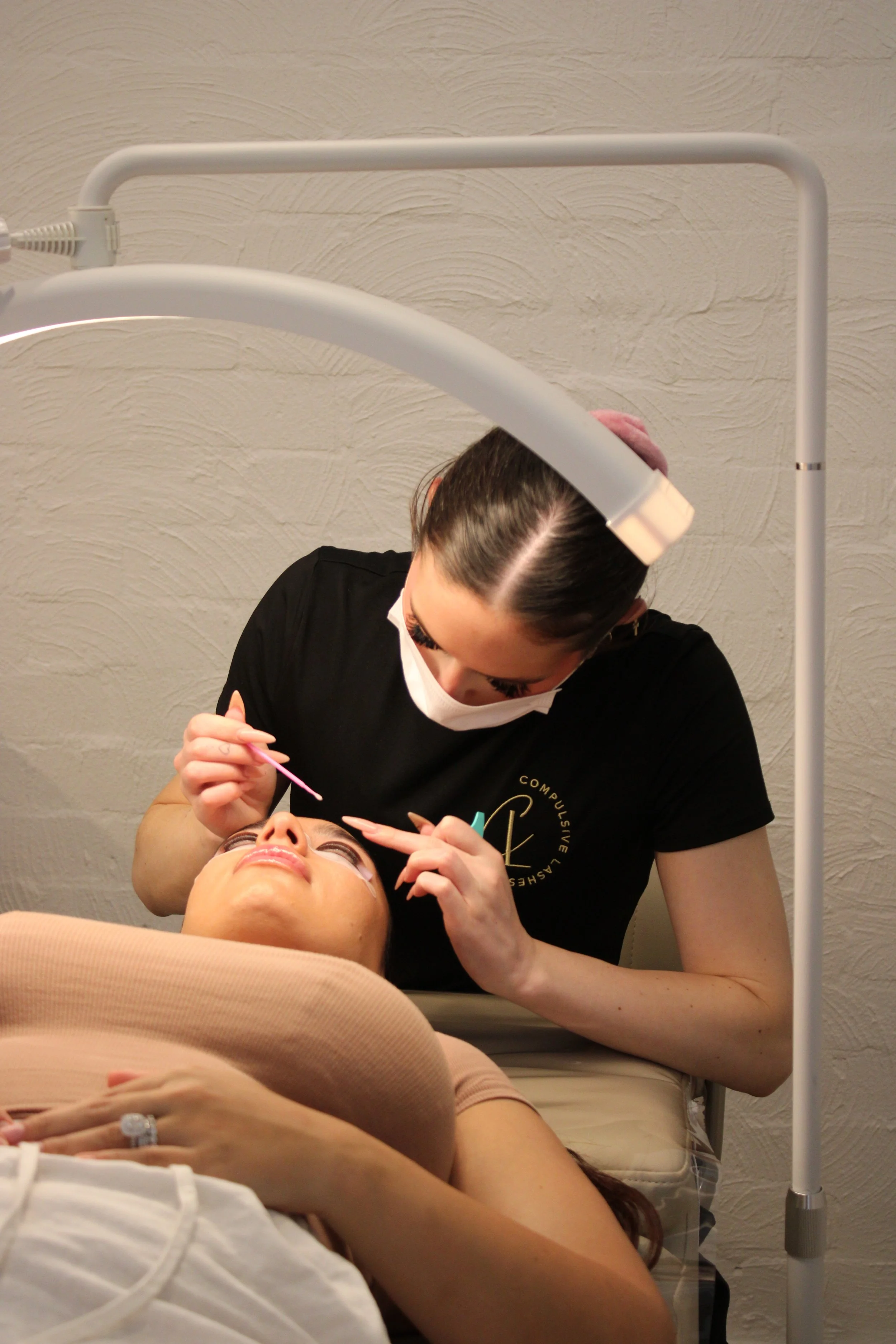 A woman is receiving a cosmetic or dental treatment from a professional in a clinical setting. The woman is lying down with eye protection, and the professional is leaning over her, holding tools while working on her face.
