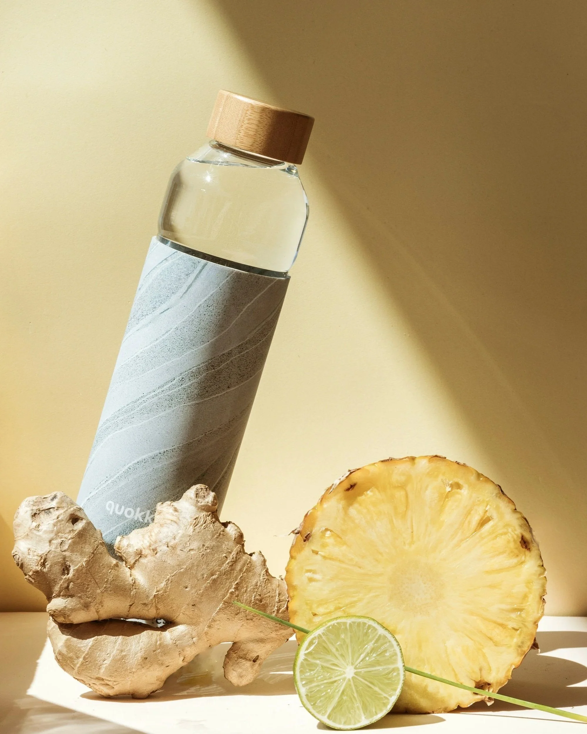 Fresh ginger root, sliced pineapple, lime slices, a bottle of water with a wooden cap, and a ceramic cup with a gray and white striped pattern placed against a yellow background.